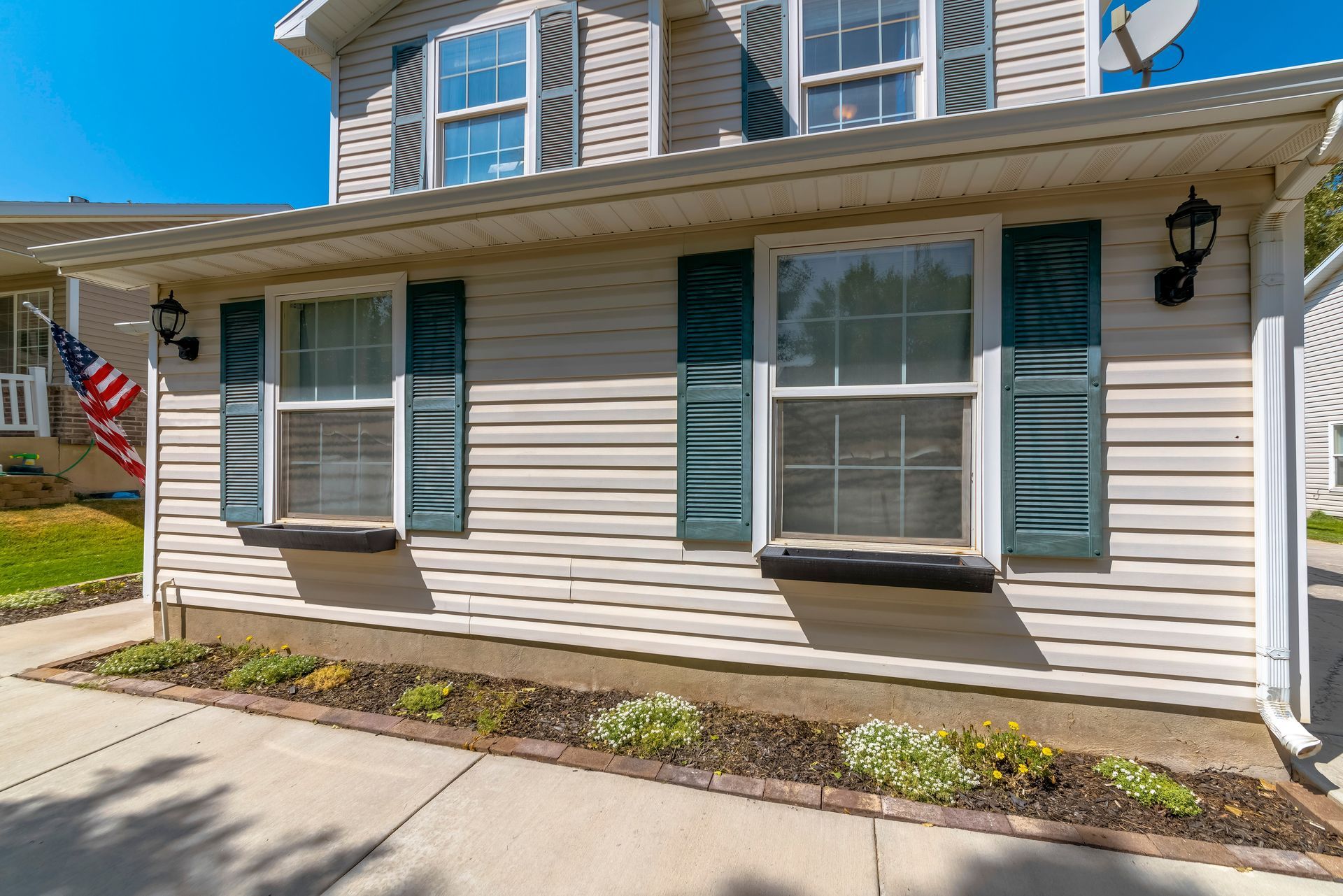 A two-story, light-colored vinyl-sided house with dark green shutters, outdoor lamps, and a small front garden bed.