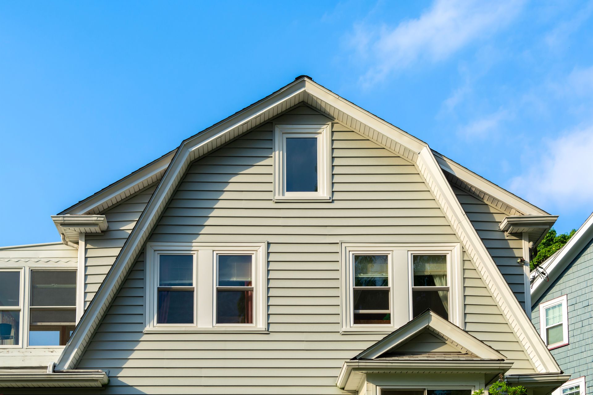 The upper section of a light-colored, vinyl-sided house featuring a gambrel roof, several windows, and a clear blue sky.