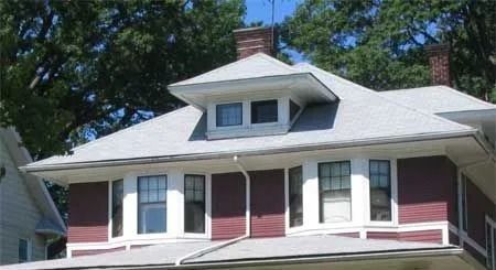 Upper story of a house with maroon siding, white trim, a gabled roof, and a centered dormer window under leafy trees.