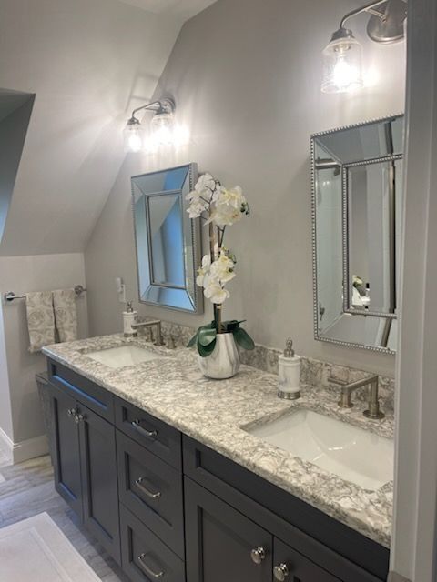 A double-sink bathroom vanity with dark gray cabinets, a light granite countertop, and two mirrors on a gray wall.