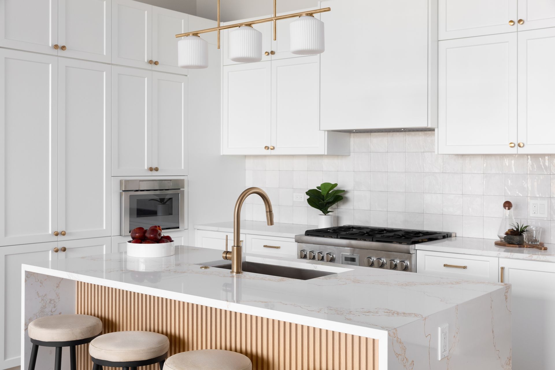 Modern white kitchen with gold hardware, a marble island with wood slat detailing, and three neutral-toned stools.
