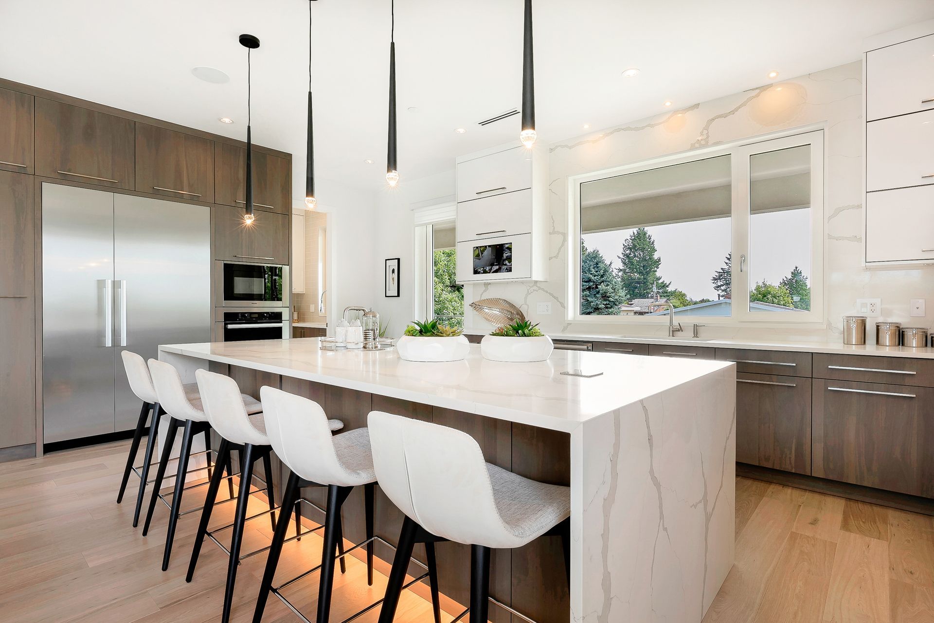 Modern kitchen with a white marble-topped island, four white bar stools, stainless steel appliances, and wood cabinets.
