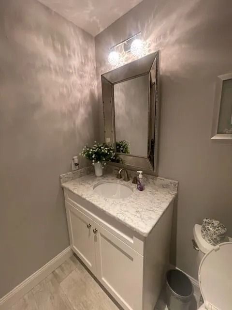 A small bathroom with a white vanity, marble countertop, large framed mirror, vanity lights, and a white toilet.