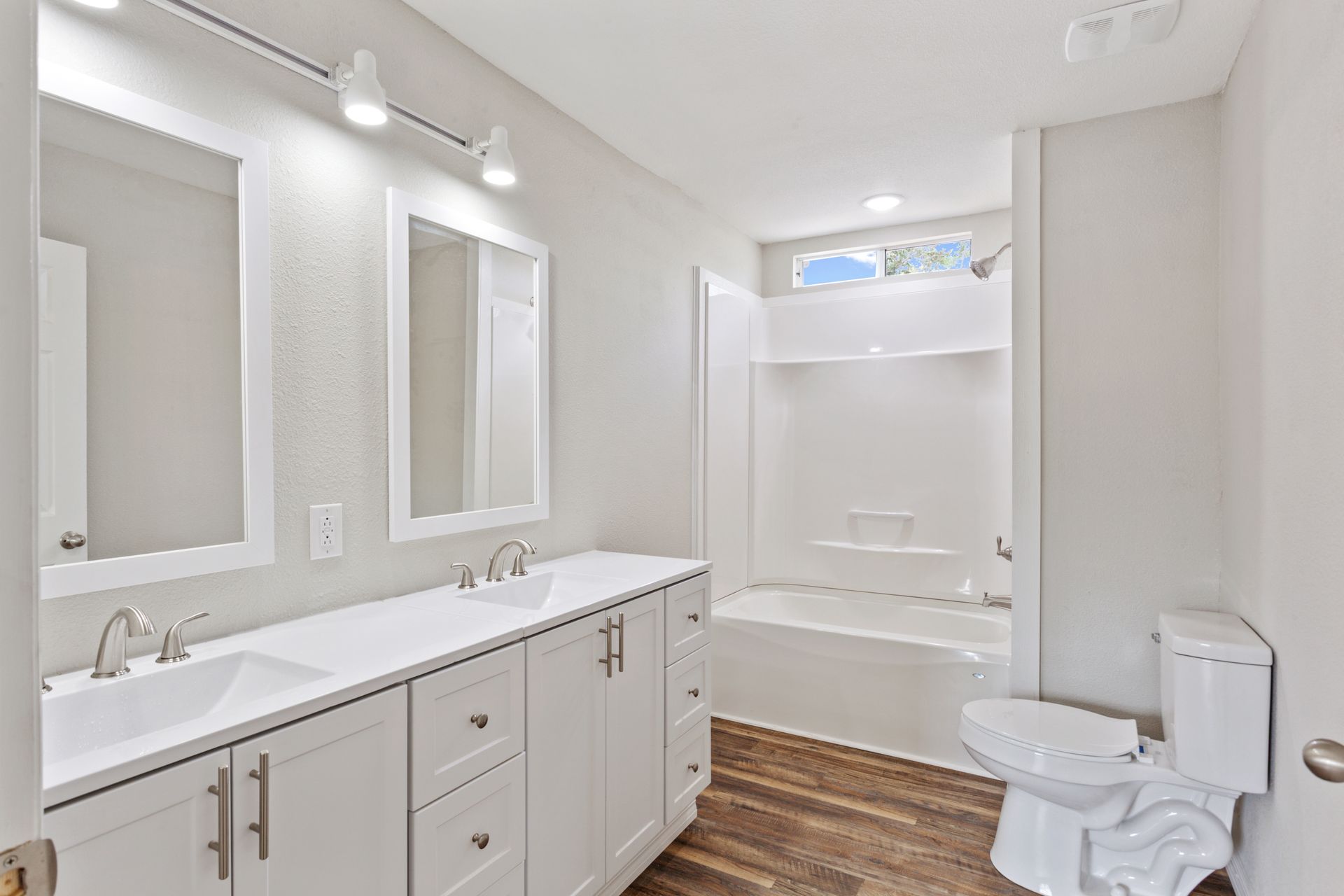 Modern bathroom with a double vanity, large mirrors, a white tub-shower combination, and wood-look flooring.