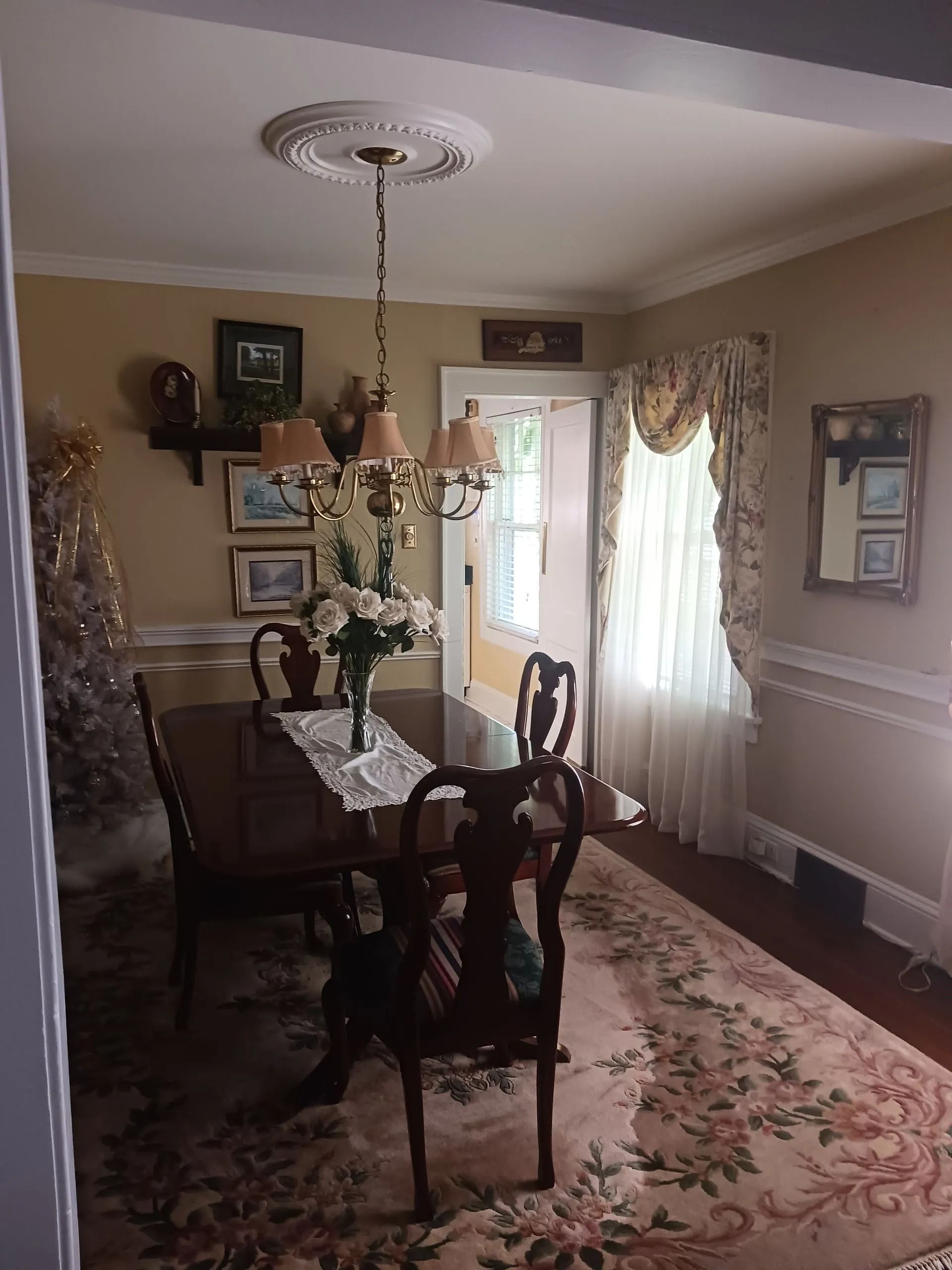A dining room featuring a wooden table, chairs, an ornate chandelier, floral rug, and curtains over a doorway.