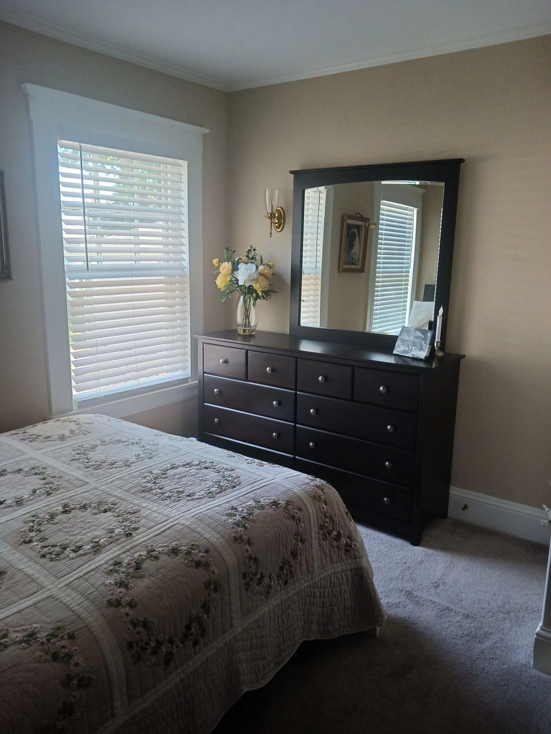 A bedroom featuring a bed with a floral quilt next to a dark wood dresser with a large mirror under a window with blinds.