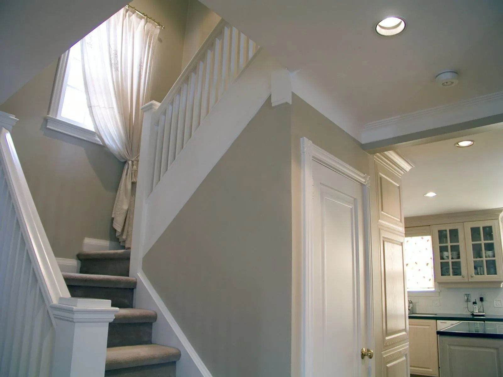 A staircase with grey carpet and white railings beside a hallway, leading to a glimpse of a kitchen with light cabinets.