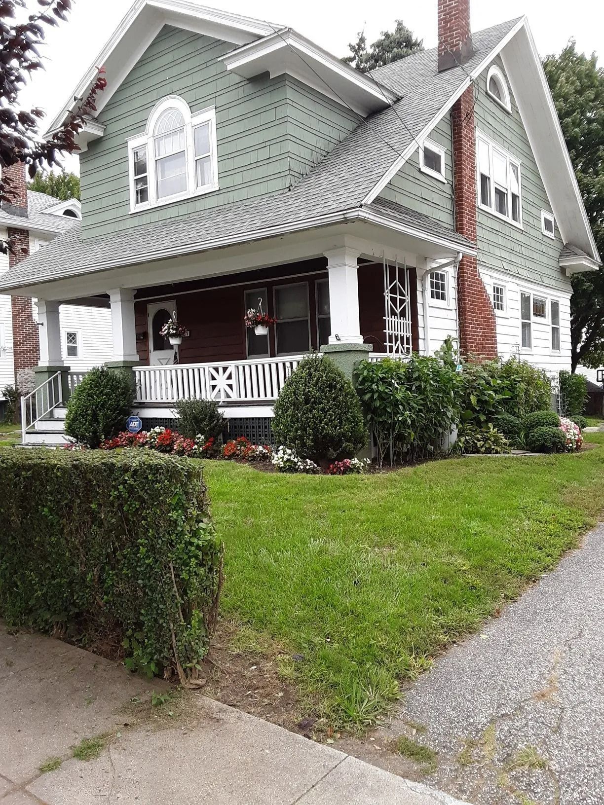 Two-story green house with a dark red ground floor, white porch railings, and a brick chimney on a grassy corner lot.