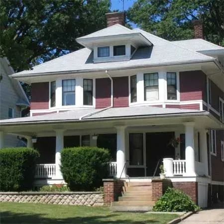 A two-story dark red house with a large front porch, white columns, and a dormer window on the roof.