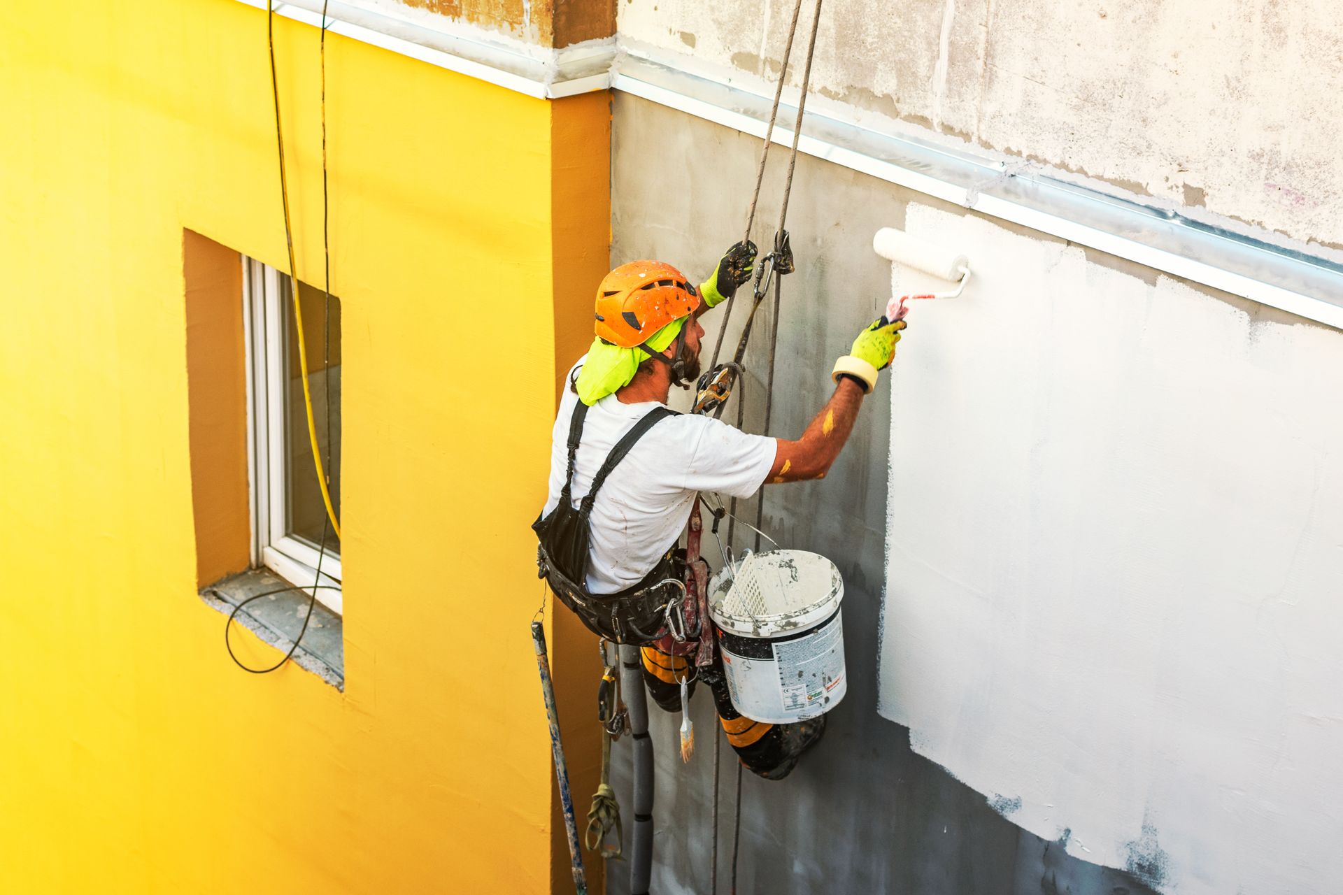 A worker in a helmet and harness rappels down the side of a building, painting a gray wall white with a paint roller.