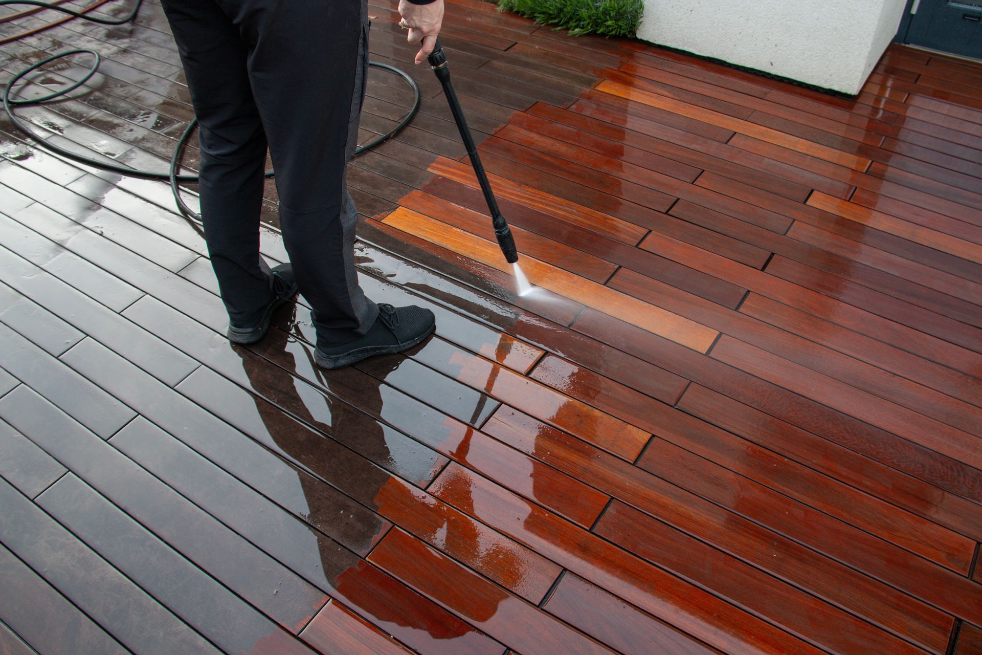 A person uses a pressure washer to clean a dark wood deck, revealing a brighter, lighter-colored surface beneath.