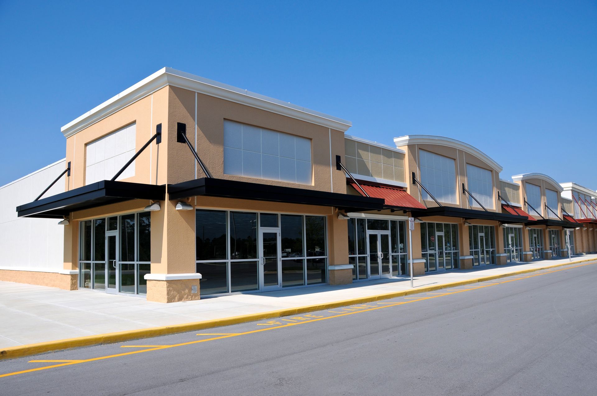 A beige strip mall with multiple empty storefronts and dark awnings under a clear blue sky.