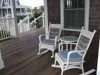 Two white wicker rocking chairs and a small round table on a wooden porch overlooking a beach house.