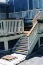Wooden stairs with white railings lead up to a raised outdoor deck on the side of a blue house.