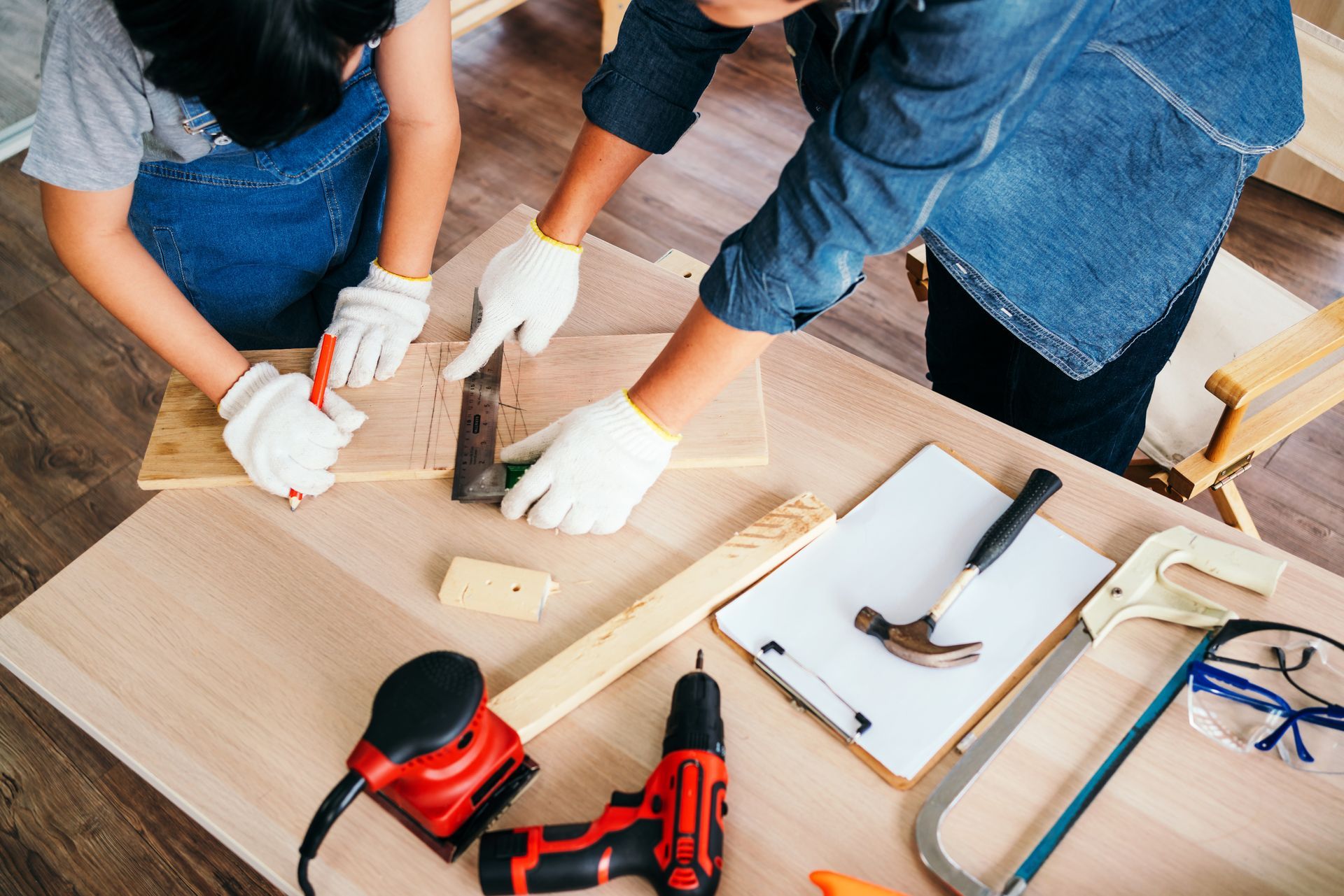 An adult and child wearing gloves use a square to measure a piece of wood on a workbench with assorted tools nearby.