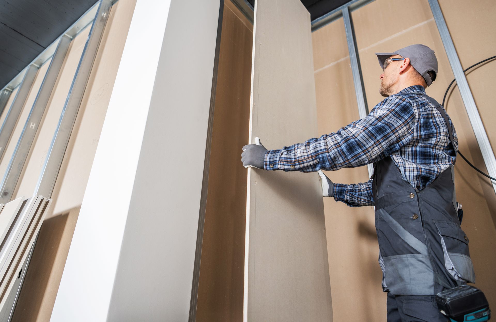 A construction worker wearing a plaid shirt and cap installs a drywall panel onto a metal wall frame.