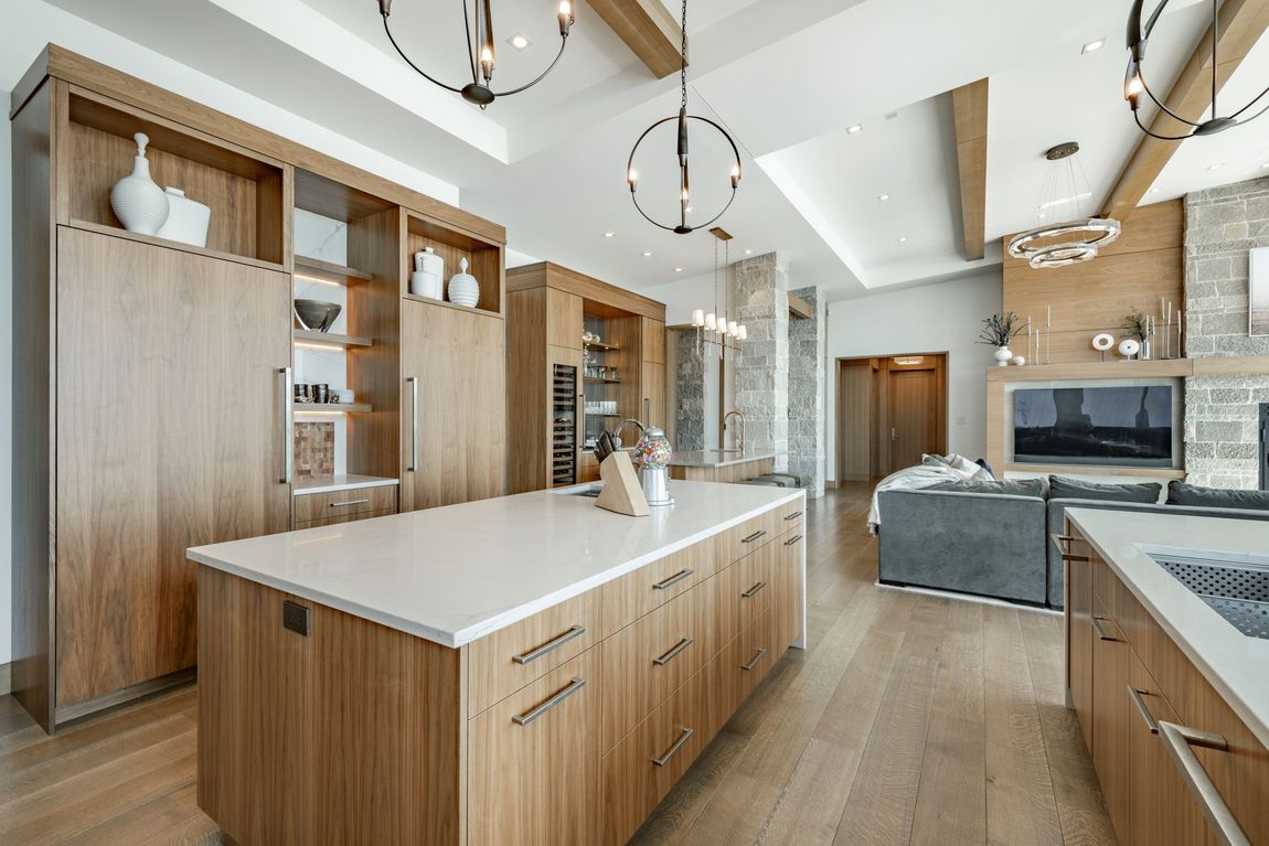Modern open-plan kitchen with light wood cabinetry, white countertops, stone fireplace, and hanging light fixtures.