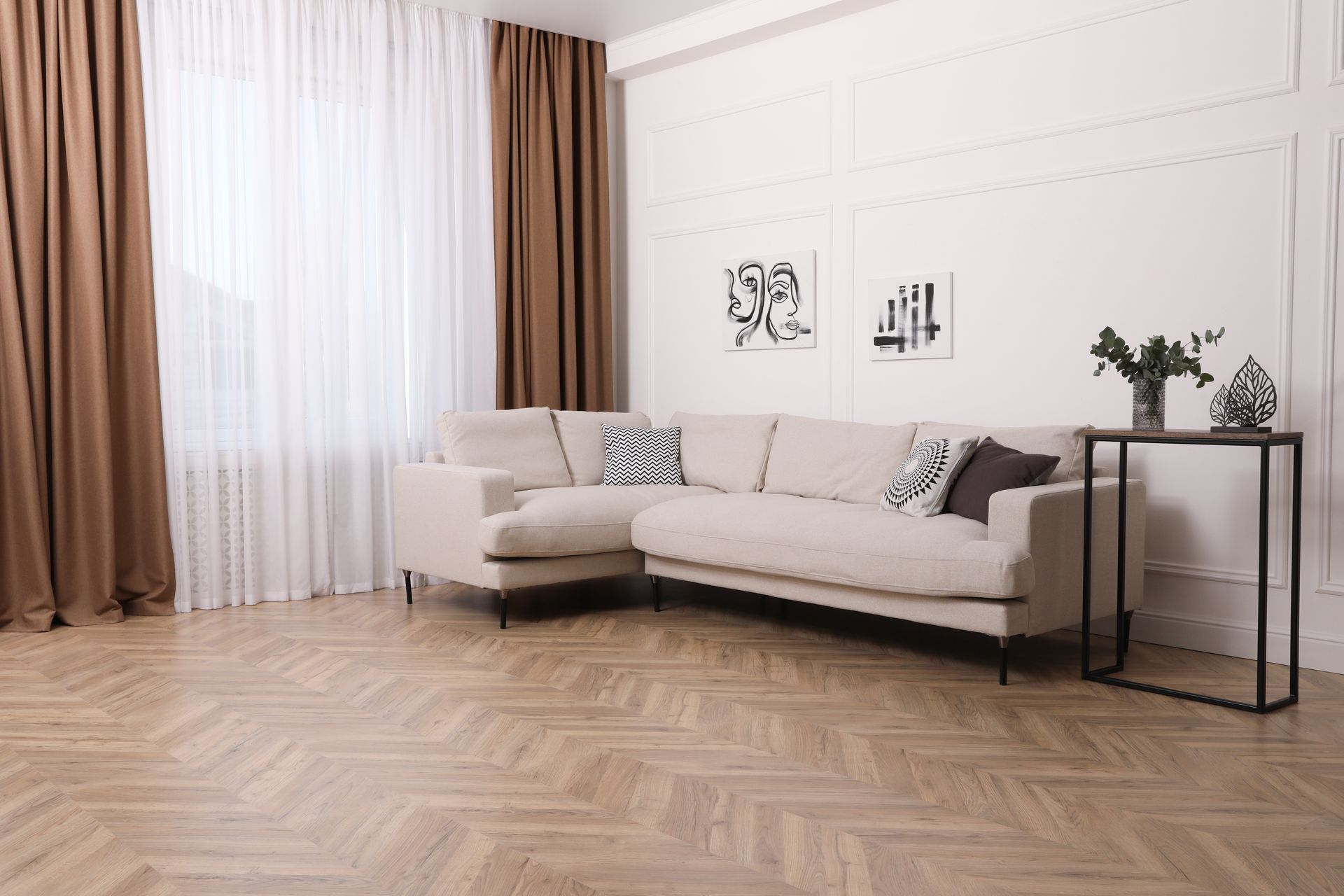 A beige sectional sofa sits in a bright living room with wood floors, brown curtains, and minimalist wall art.