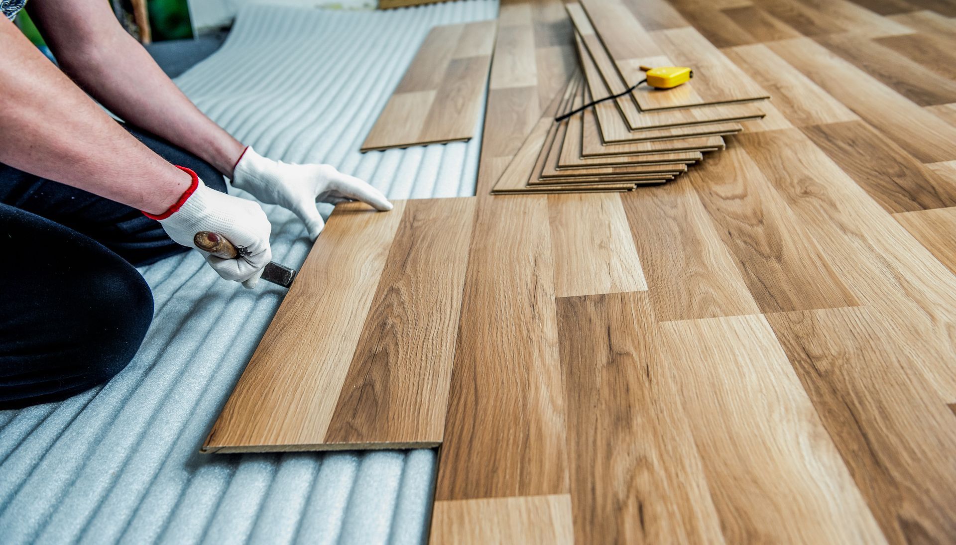 A person wearing gloves cuts a wood-look laminate floor plank with a utility knife over white foam underlayment.