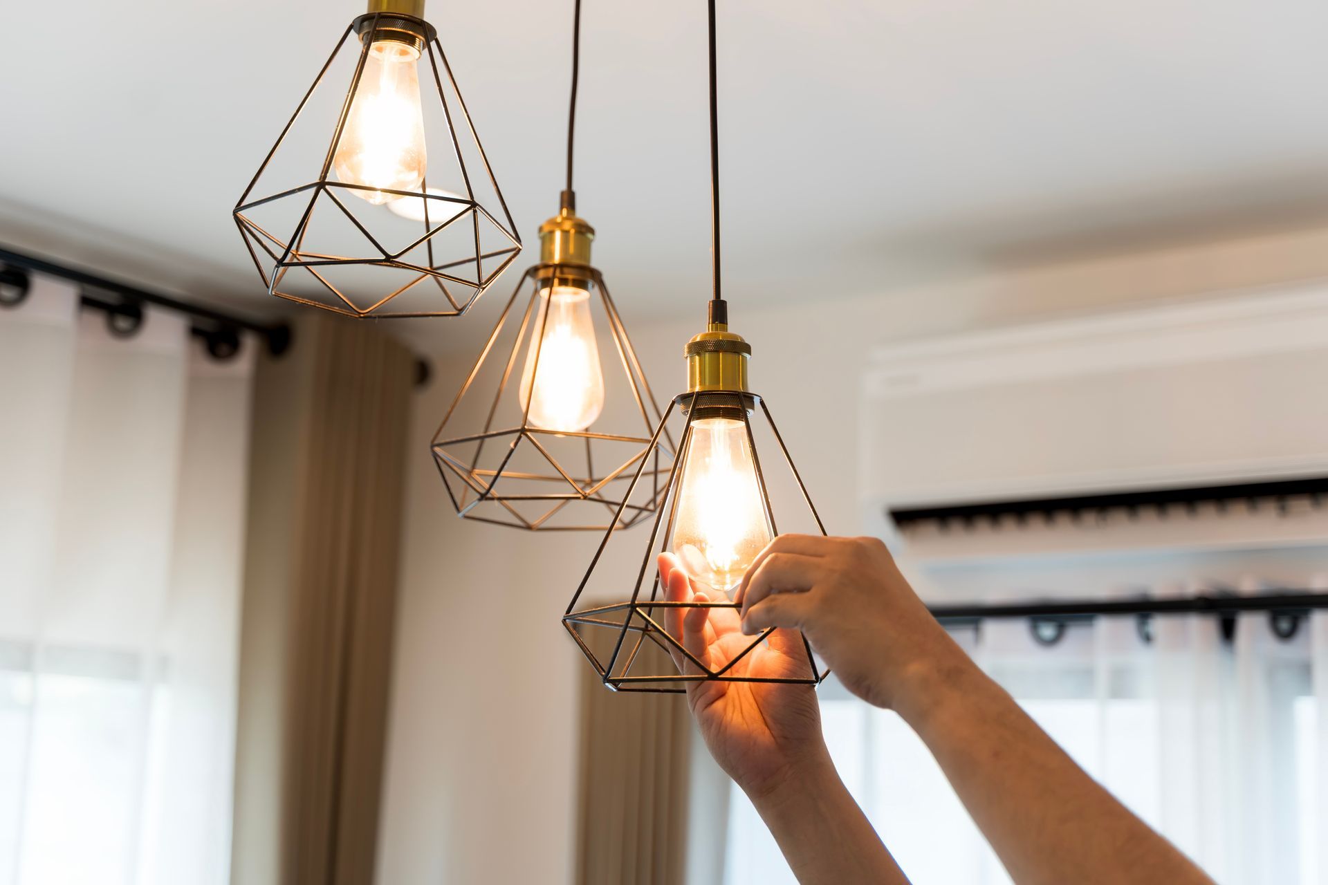 A person adjusts a glowing geometric pendant light in a room with a white curtain and wall-mounted air conditioner.