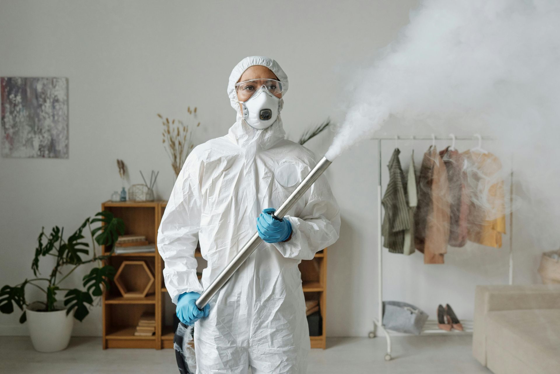 Person in blue suit, mask, and backpack vacuuming wooden floor in a room with a window.