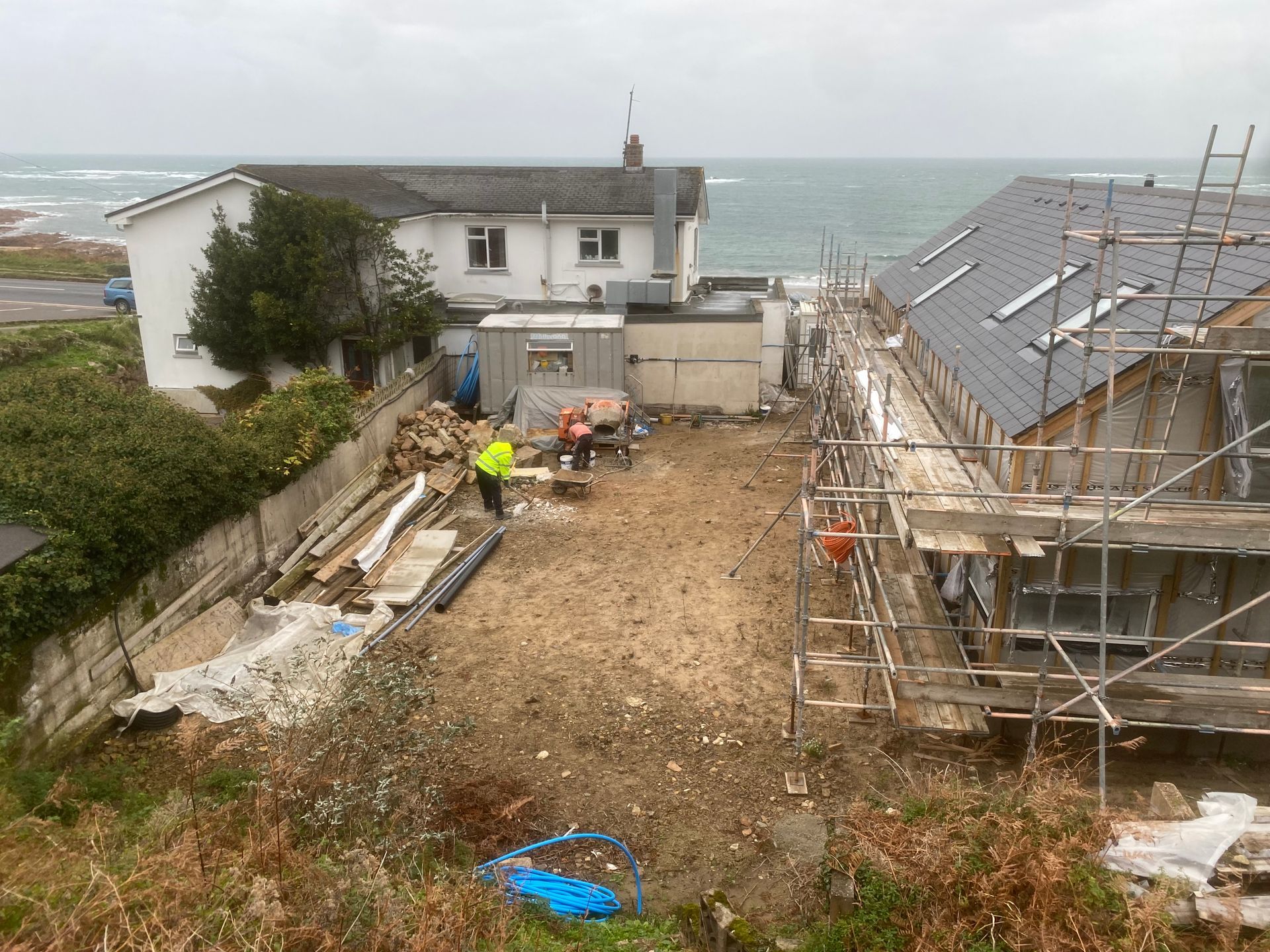 Construction site with two houses, one being built, ocean in the background. Two workers are present.