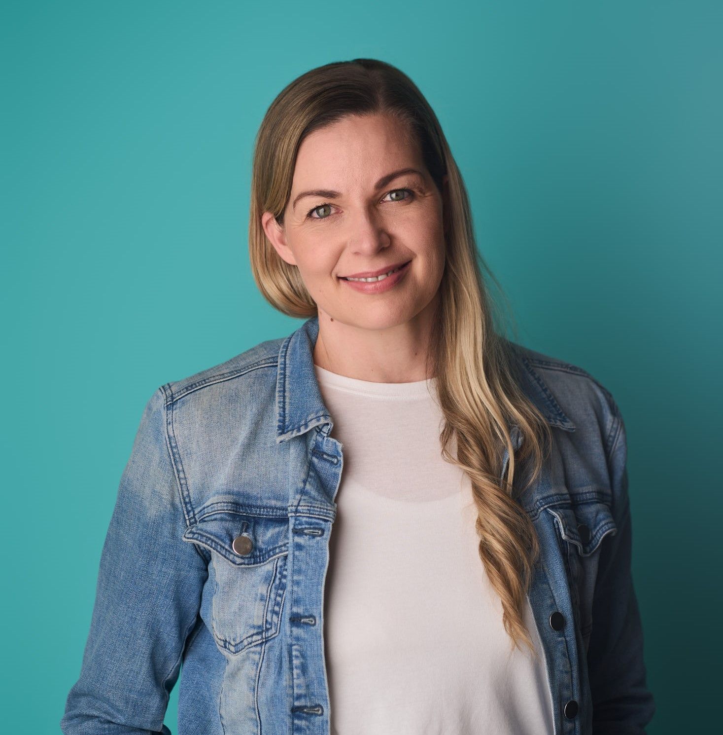 A woman wearing a denim jacket and a white shirt is standing in front of a blue background.
