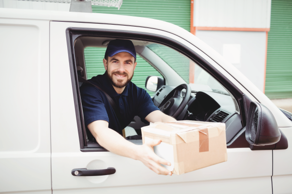 A delivery driver in a navy uniform smiles from the driver's seat of a white van, holding out a cardboard package.