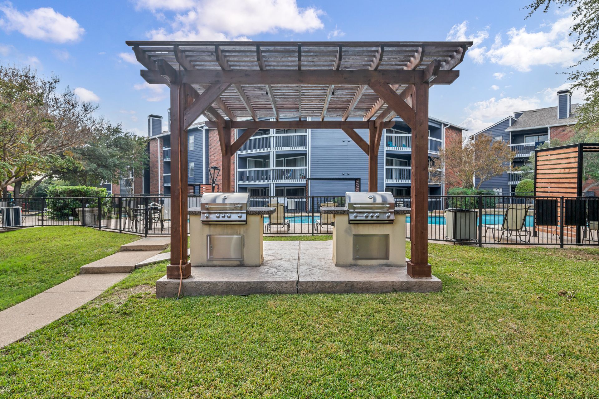 A pergola with two grills underneath it in a grassy area.