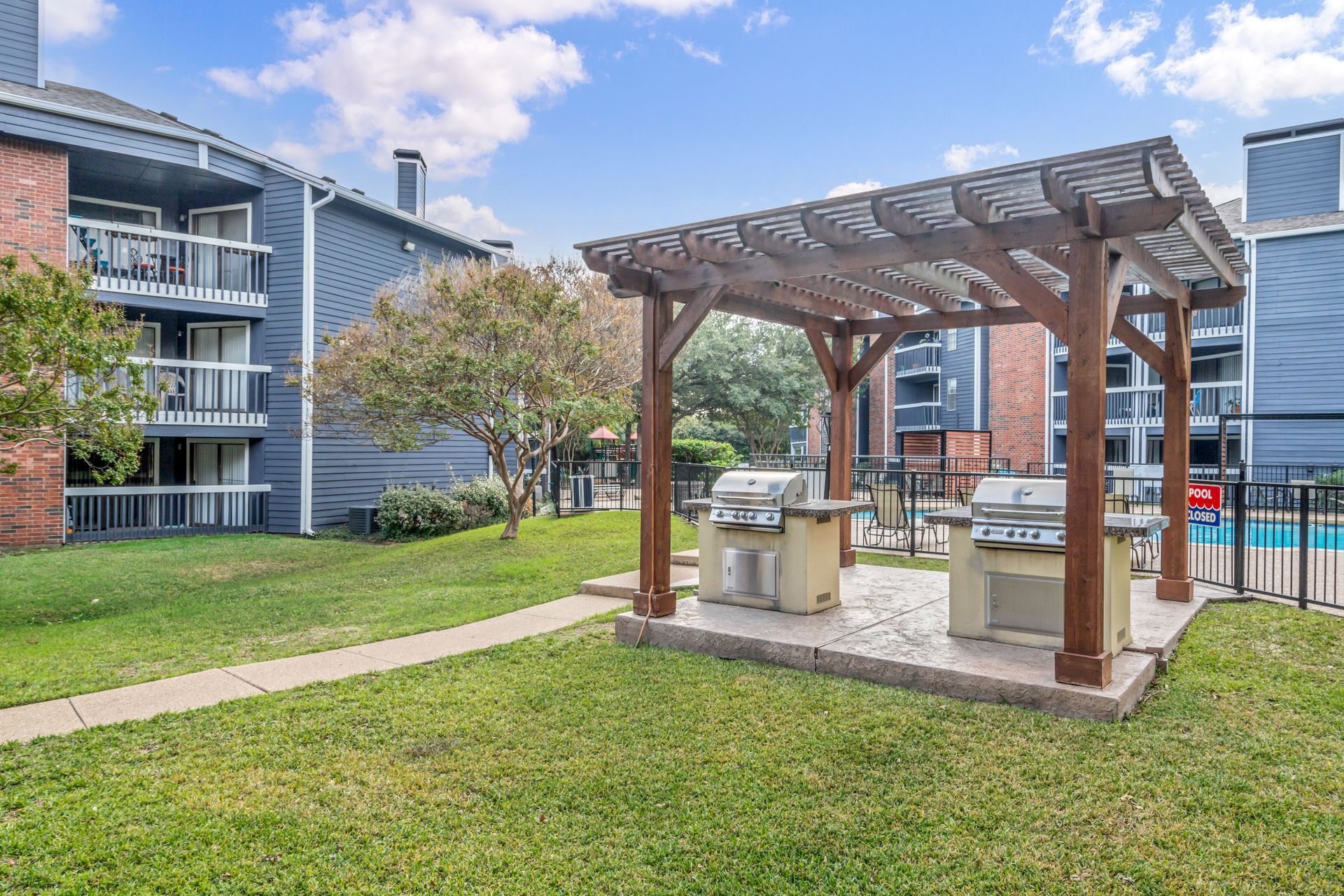 There is a grill under a pergola in the middle of a lush green field.