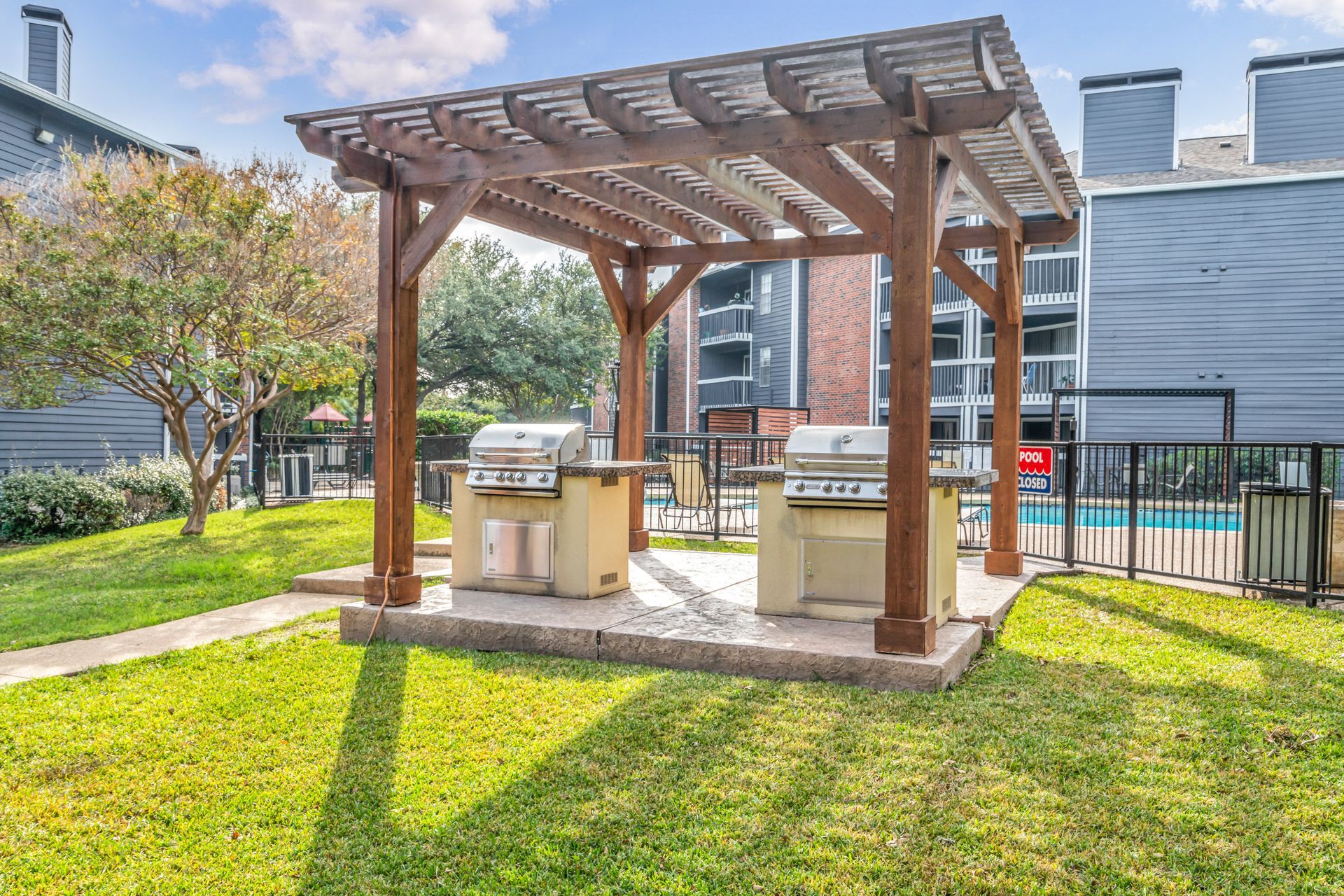 There is a grill under a pergola in the middle of a lush green field.