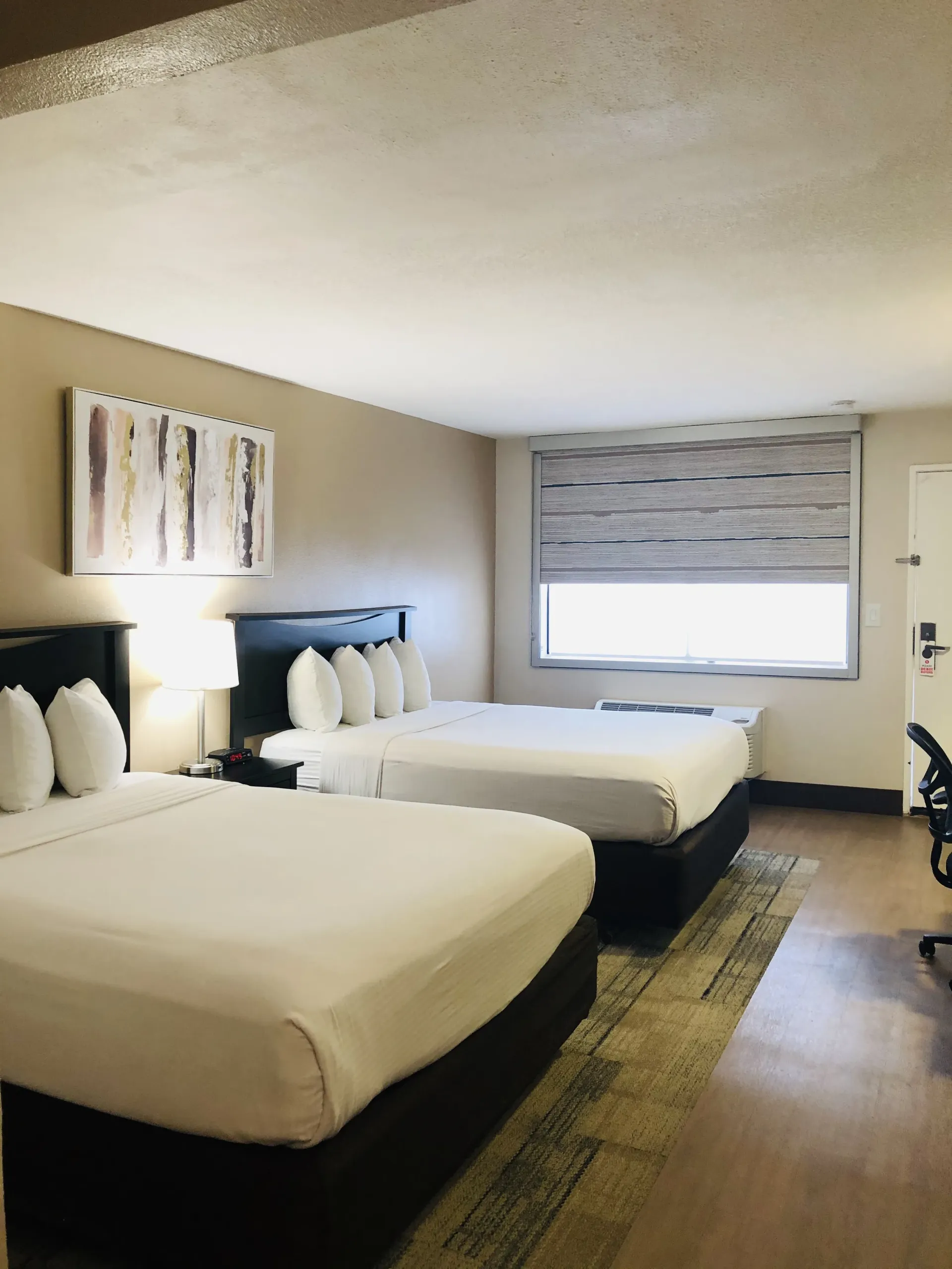 Hotel room with two neatly made beds, beige walls, window with blinds, and bedside lamp illuminated