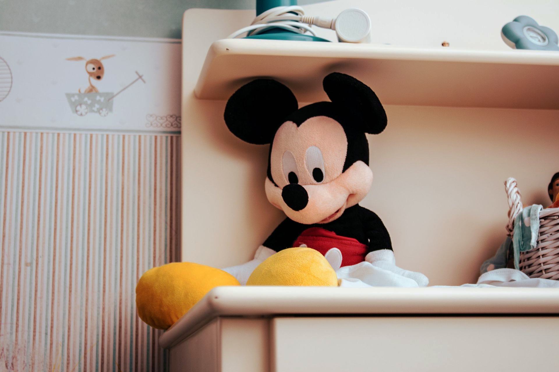 Mickey Mouse plush seated on a desk shelf in a bedroom, with yellow shoes and red shorts