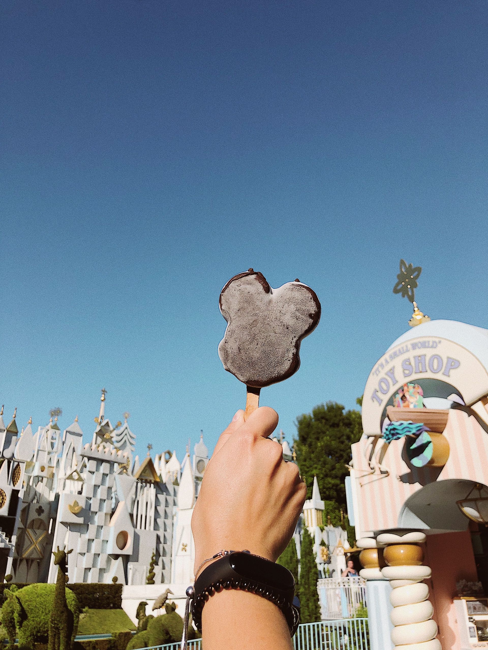 Hand holding a Mickey Mouse-shaped ice cream cone against a clear blue sky at an amusement park