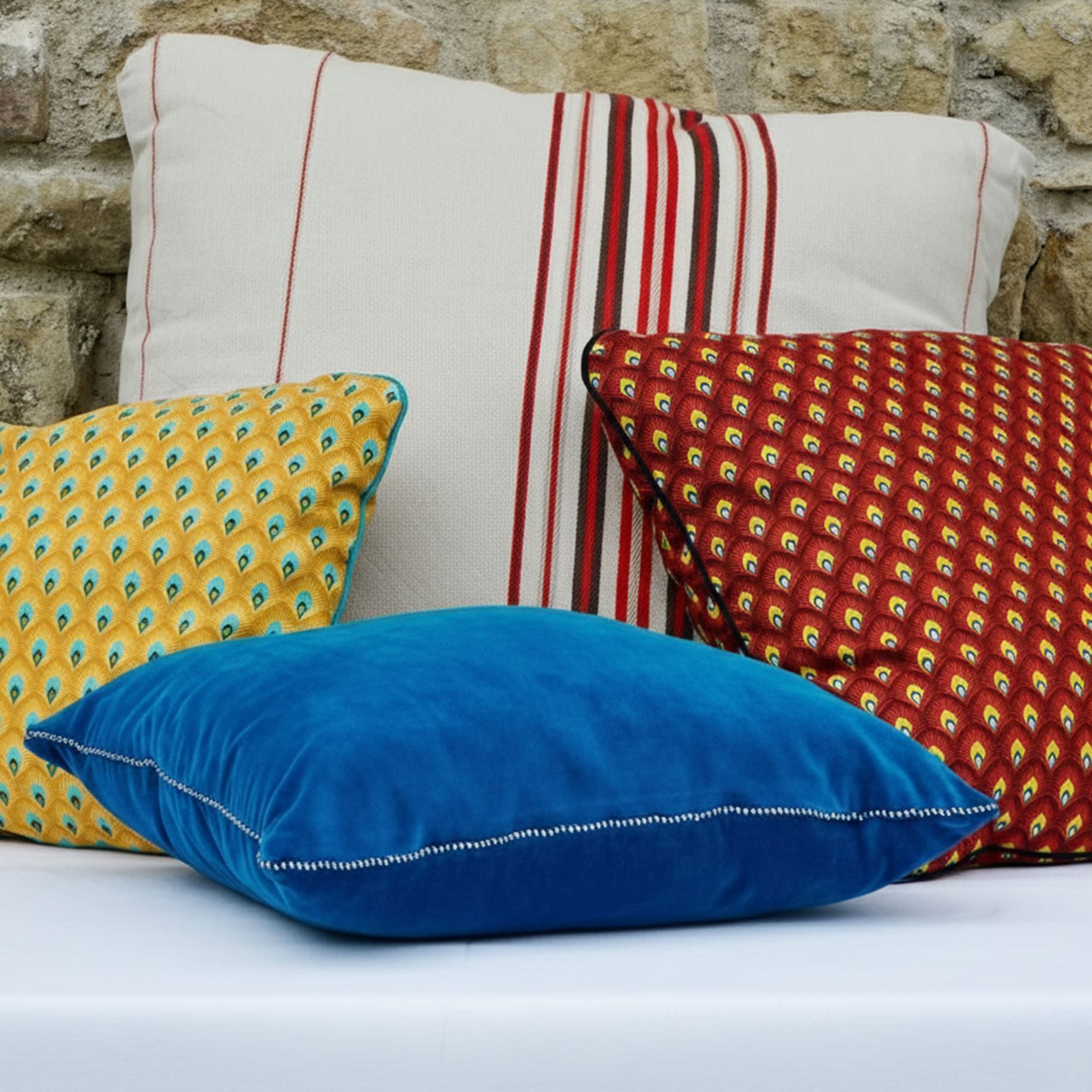 Coussins décoratifs TOKILIA uniques et écoresponsables : velours bleu, jaune, rouge et rayé rouge/blanc sur mur en pierre.