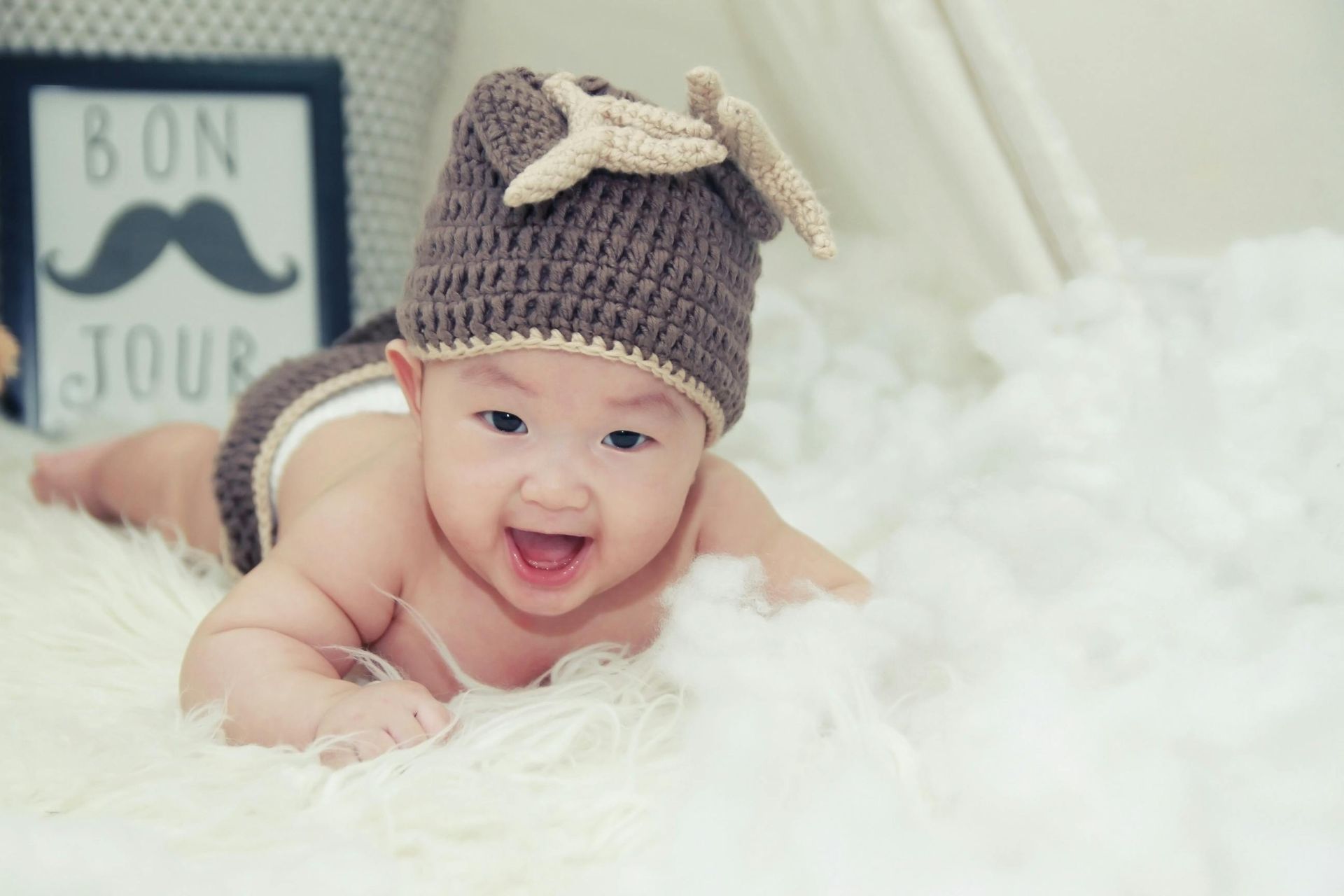 Bébé souriant, coiffé d'un chapeau à cornes marron et portant une couche, allongé sur de la fourrure blanche. Panneau « Bonjour » en arrière-plan.