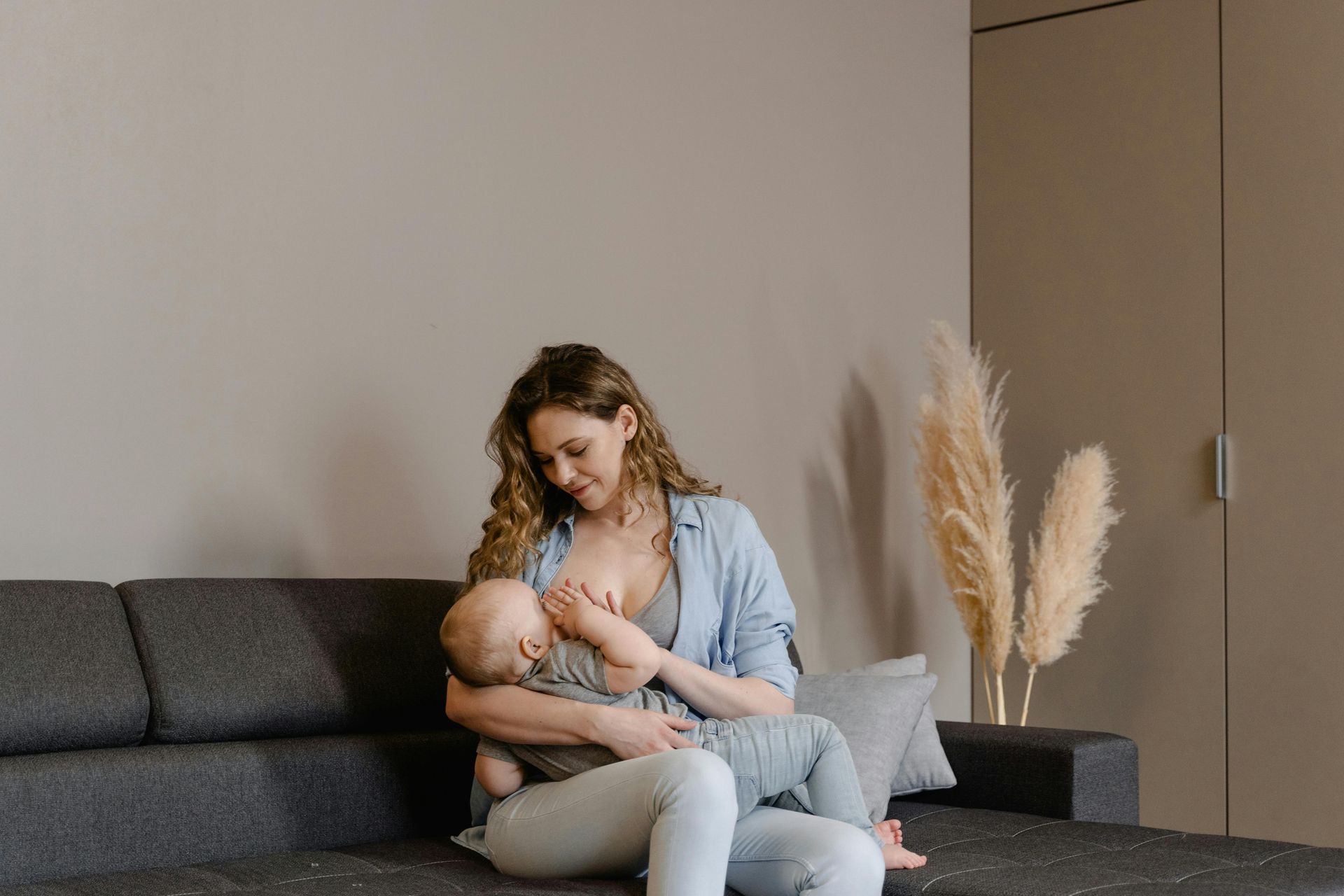 Une femme allaite son bébé sur un canapé gris dans une pièce moderne agrémentée de plantes décoratives.