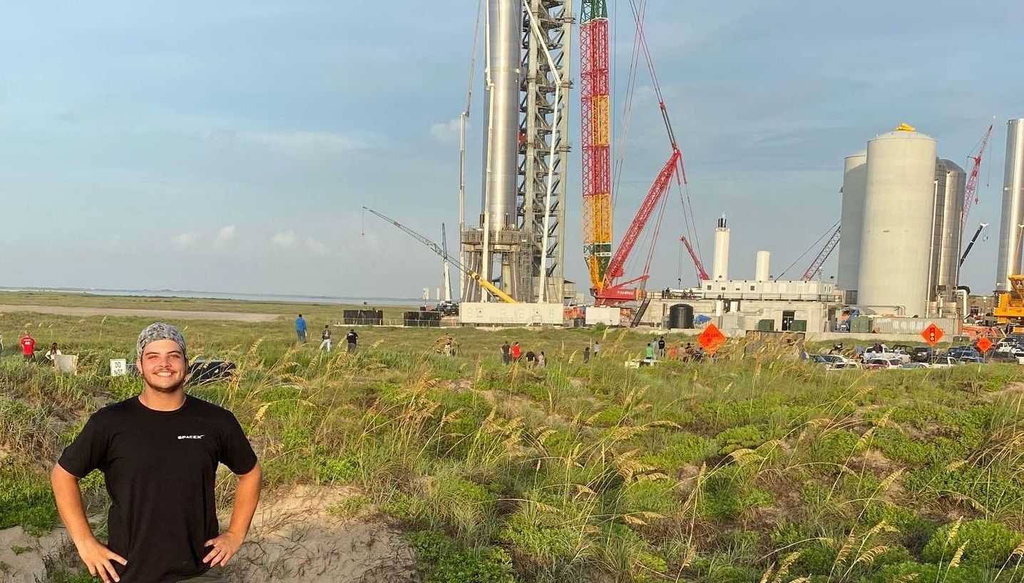 A person stands in a grassy field in front of a SpaceX launch tower and large industrial tanks under a clear sky.