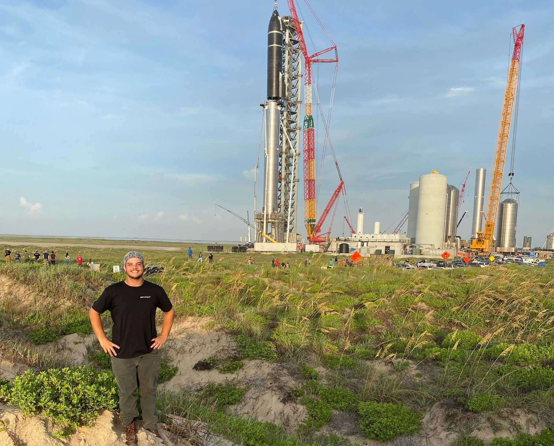A person stands in a grassy field in front of a SpaceX Starship launch tower and rocket under a clear blue sky.