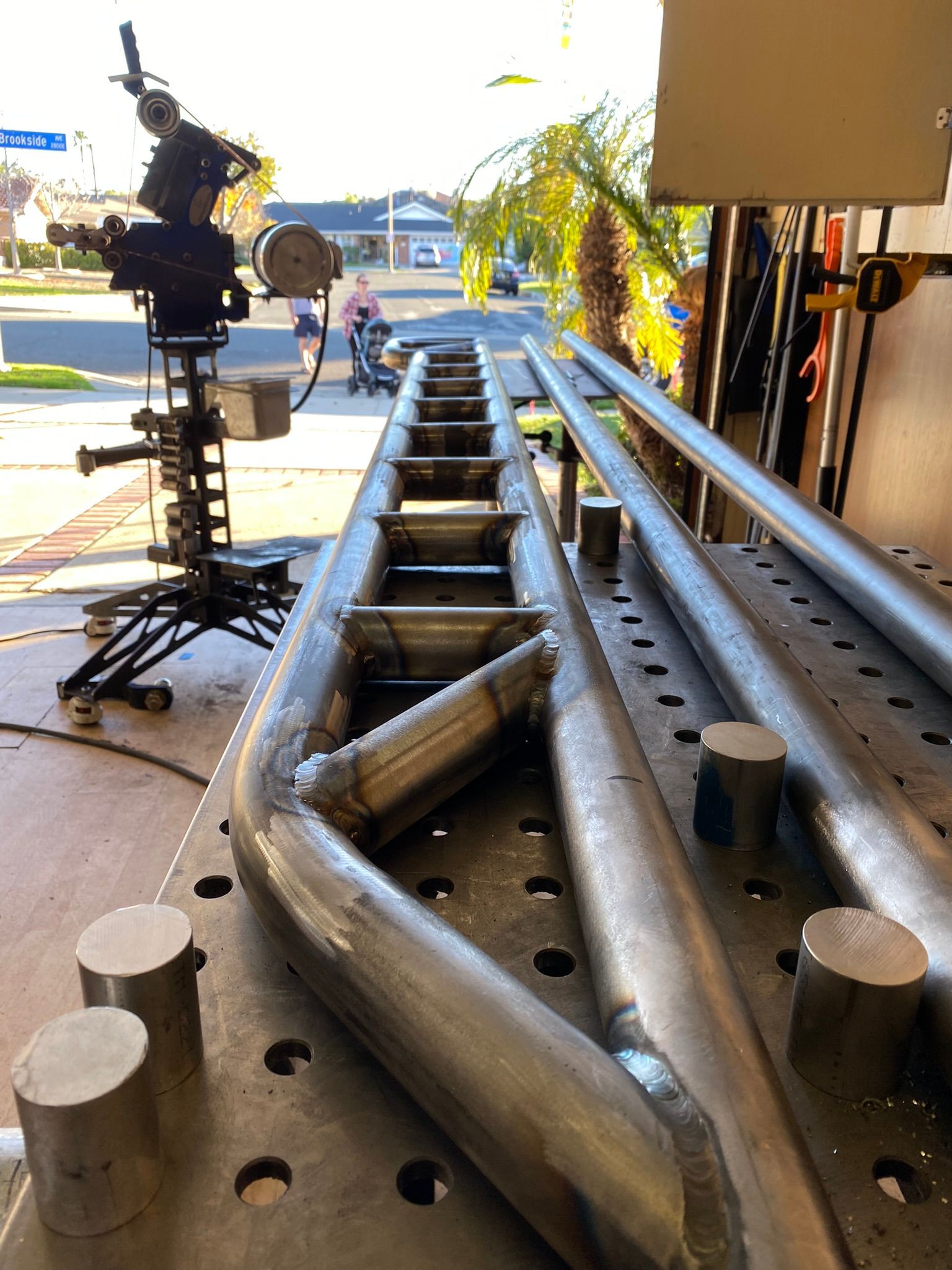 A metal structure being fabricated on a welding table in a garage with a camera rig visible in the background.