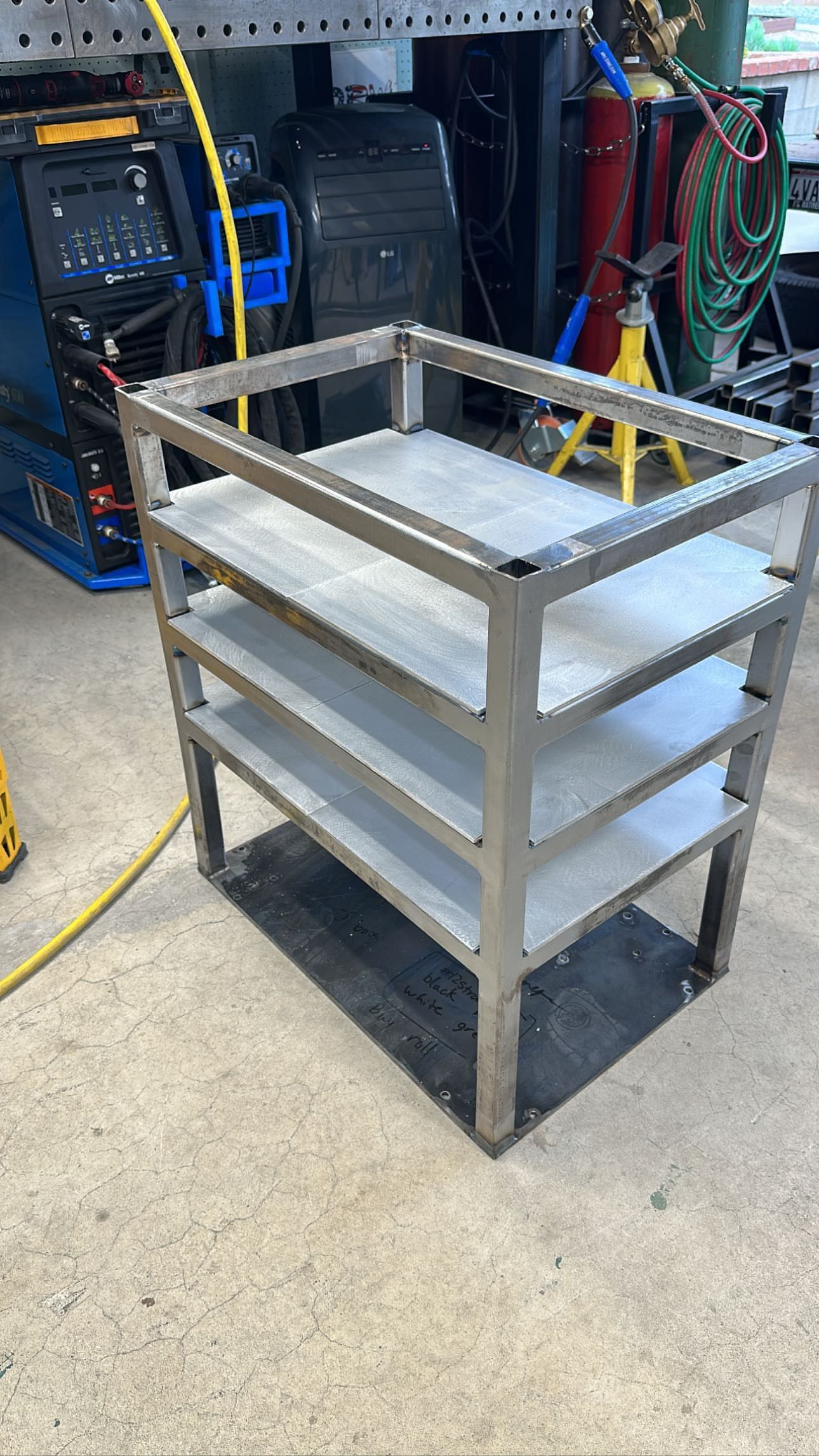 A metal welding cart with three shelves sitting on a concrete floor in a workshop.