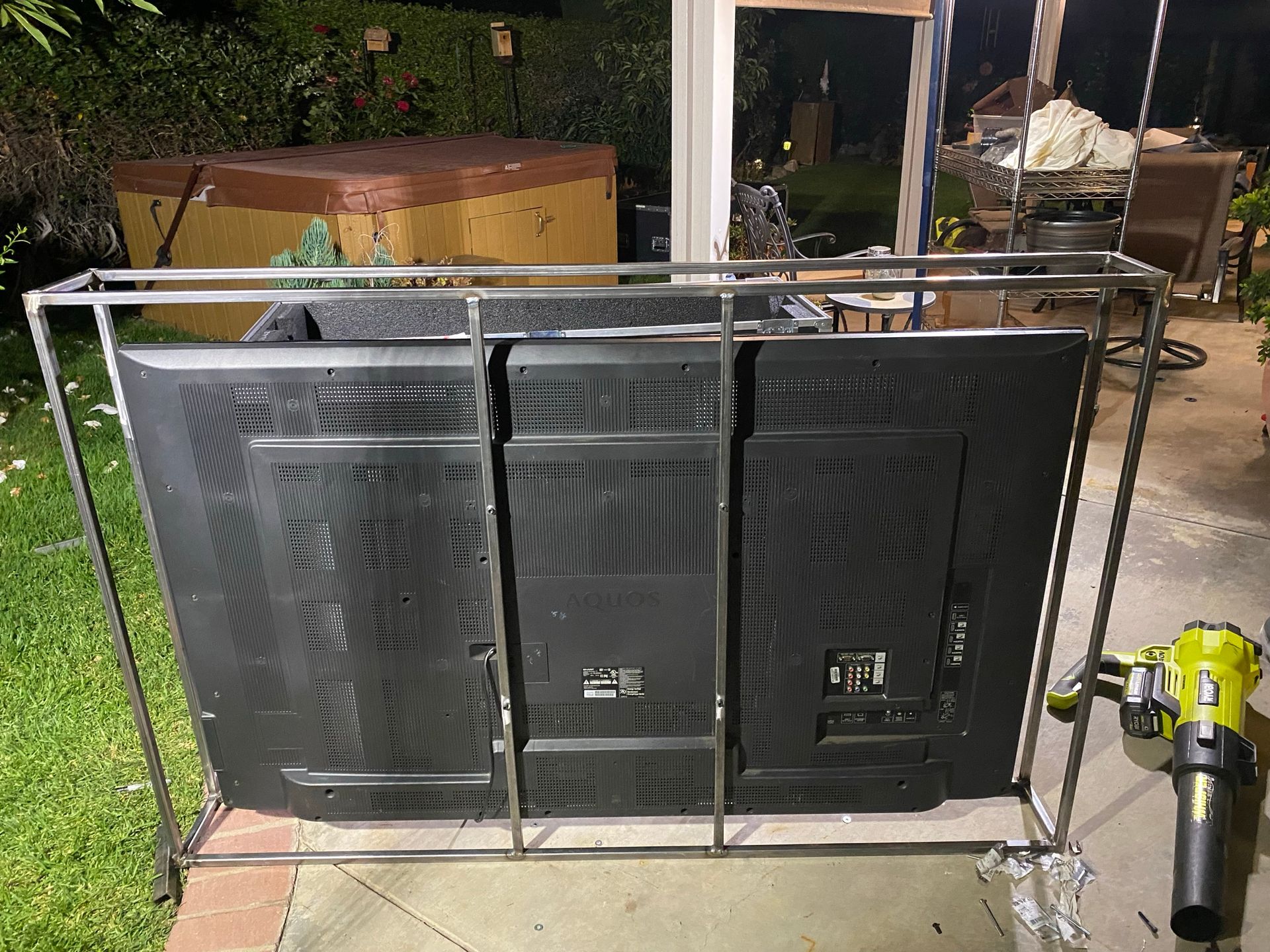 Three flat-screen TVs mounted side-by-side on a metal frame, standing outdoors on a patio next to a leaf blower.