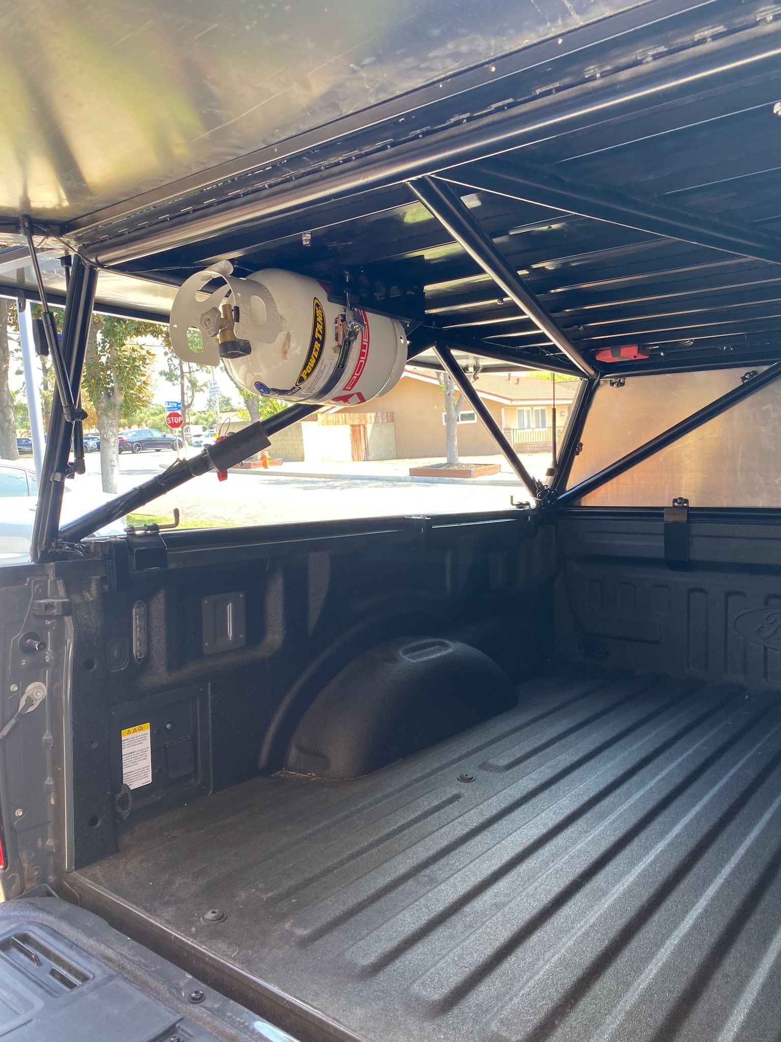 A white propane tank strapped horizontally to the black metal rack inside the bed of a pickup truck.