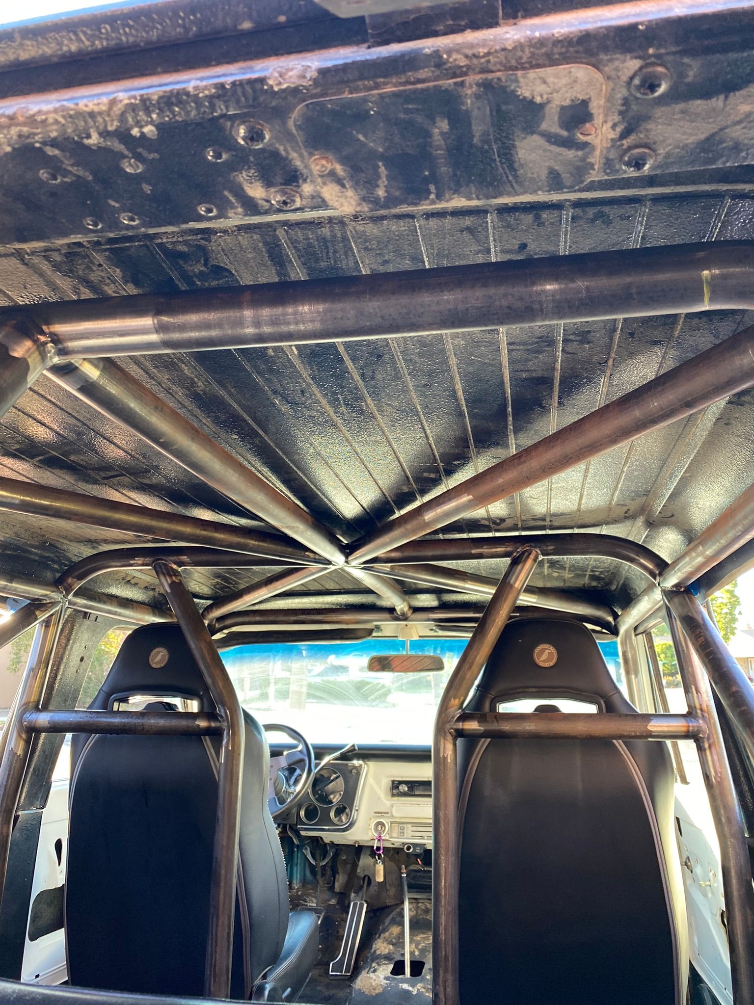 Interior view of a vehicle featuring a complex metal roll cage structure, black racing seats, and a metallic roof.