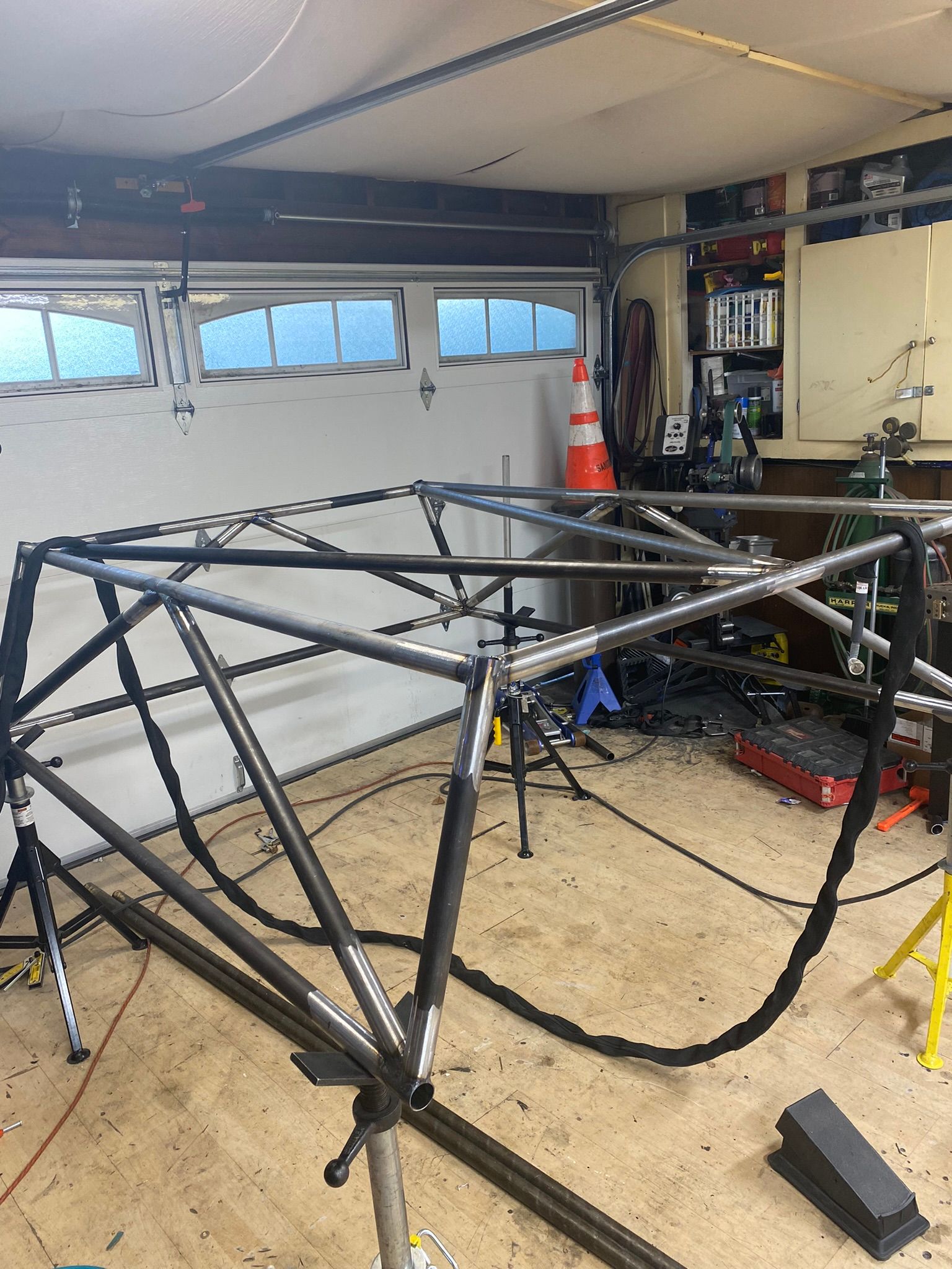 A metal roll cage frame for a vehicle under construction, positioned on stands inside a garage workshop.