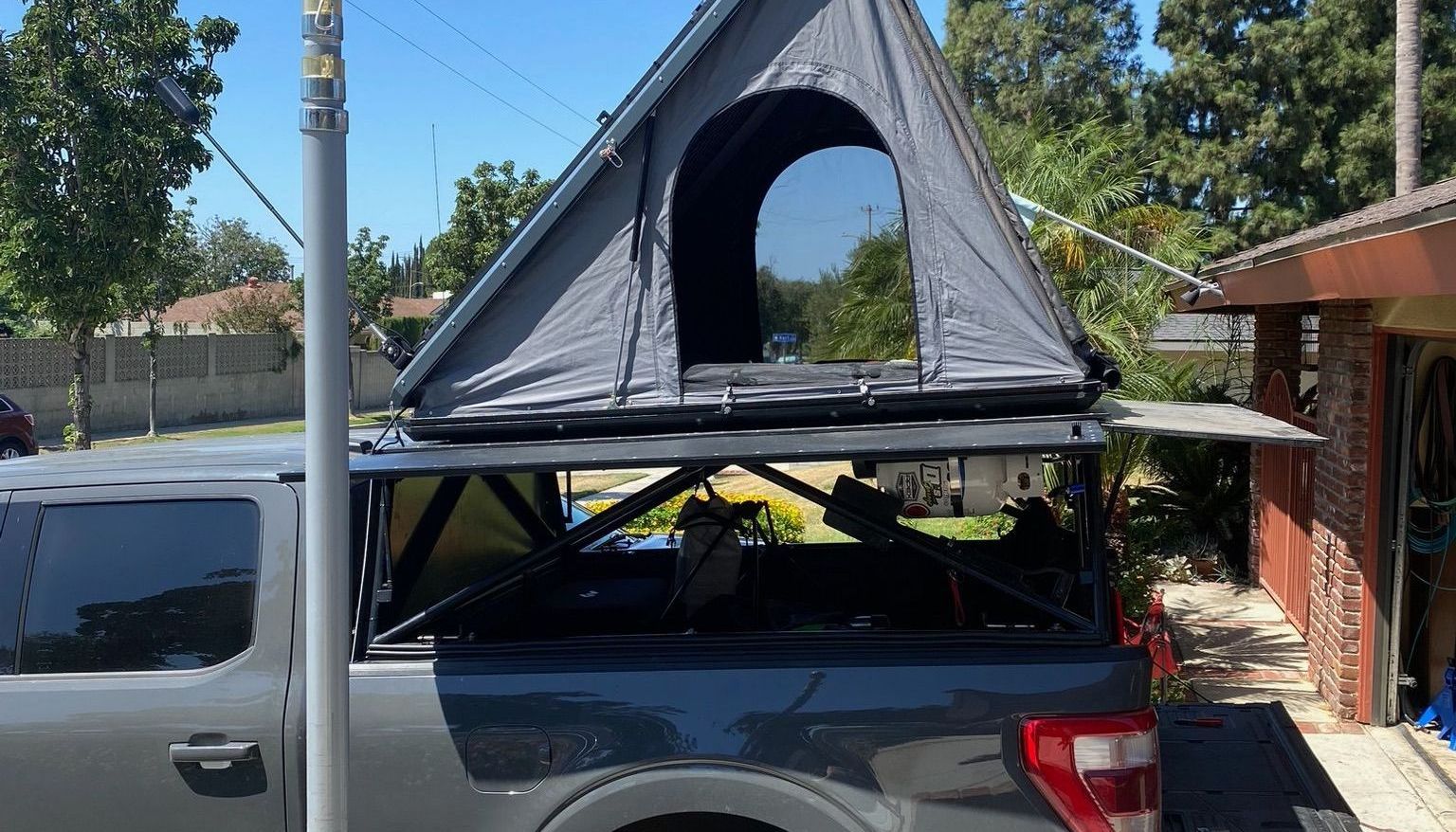 A grey truck parked outdoors with a triangular, dark grey rooftop tent mounted on a black bed rack.