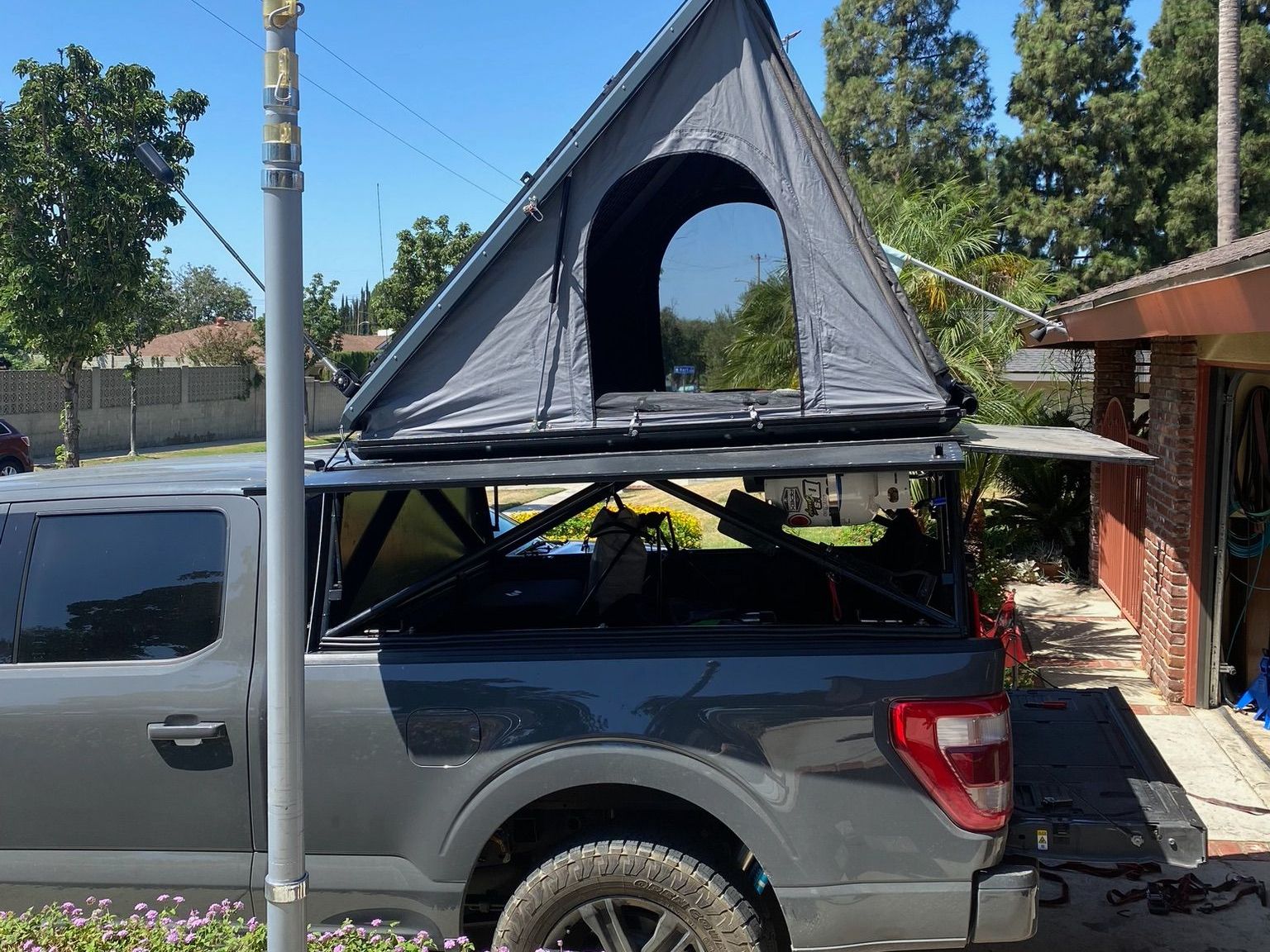 A gray pickup truck parked in a driveway with a dark triangular rooftop tent set up on a metal bed rack.