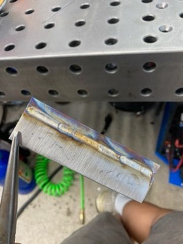 A metal rectangular piece held by pliers, displaying a TIG-welded seam with heat-tinted edges on a workbench.