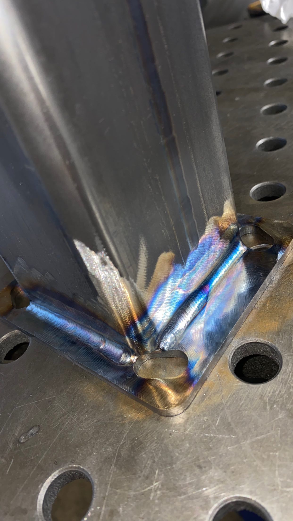 A close-up view of a metal square tube welded to a flat steel base plate, showing heat-tinted blue steel at the joints.