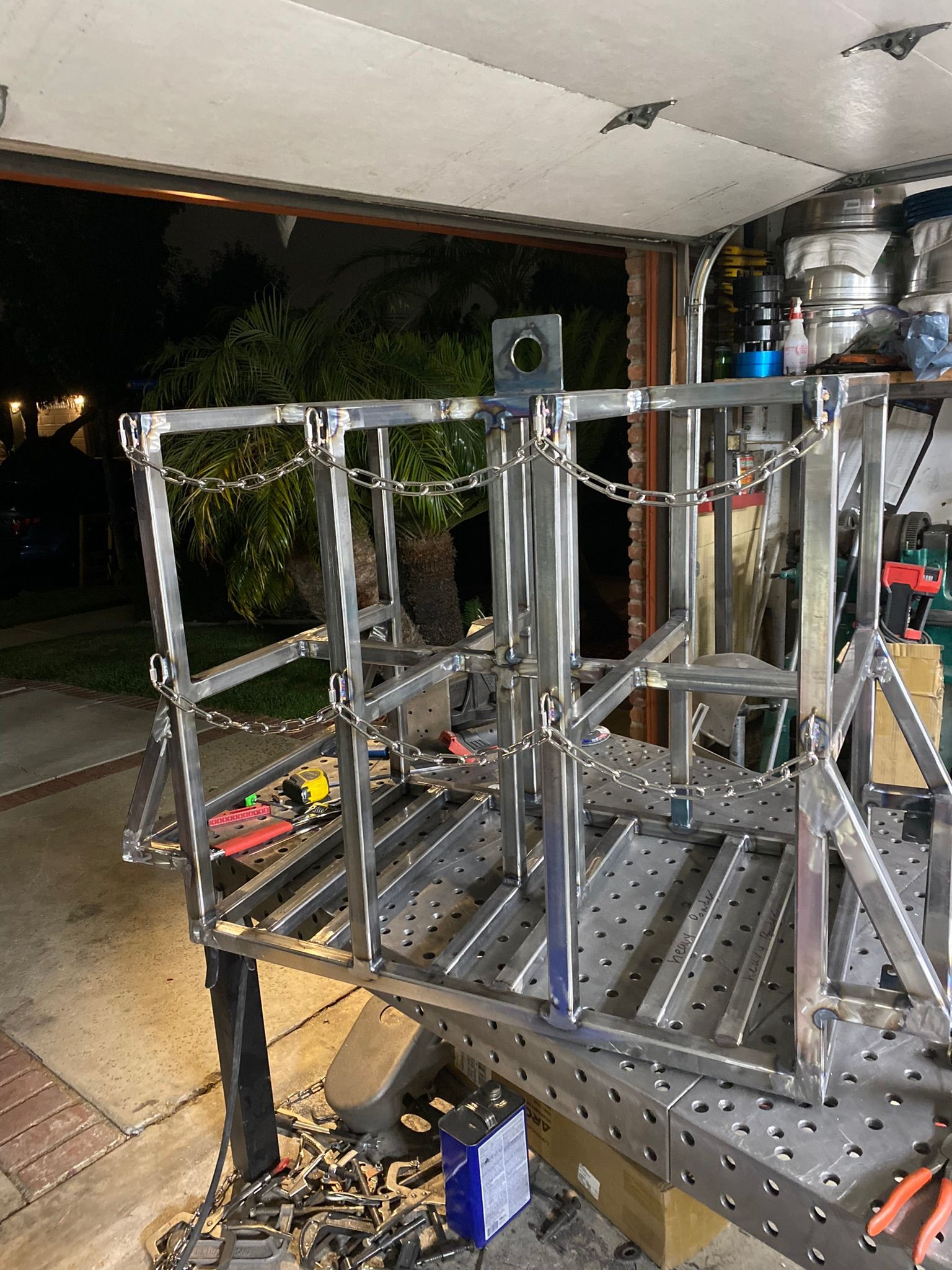 A welded metal frame structure, likely an equipment cage, sits on a perforated work table in a garage workshop.